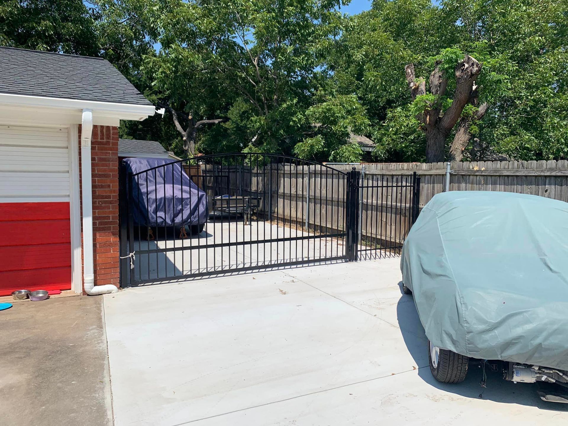 Black iron gate in a concrete driveway, flanked by two car covers. Garage on the left, trees in the background.