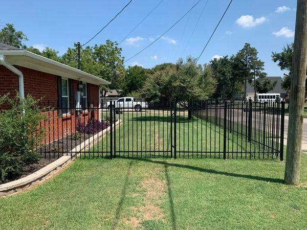 Black metal fence surrounds a green lawn, in front of a brick house, under a blue sky.