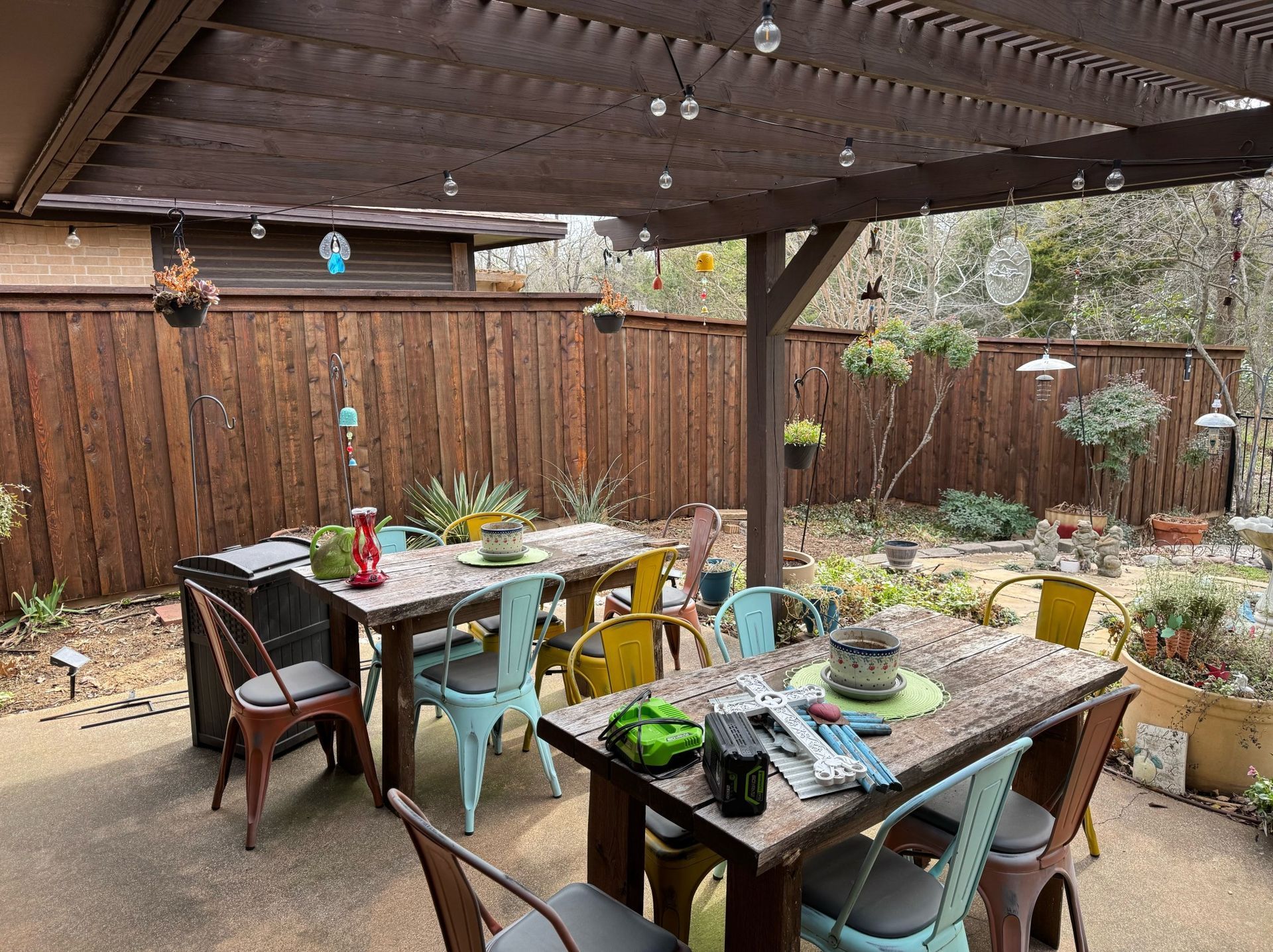 Outdoor dining area with tables and colorful chairs under a wooden pergola. Wooden fence and hanging plants surround the space.