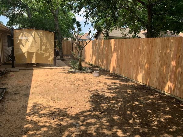 A backyard with a new wooden fence, dirt ground, and shade from trees.