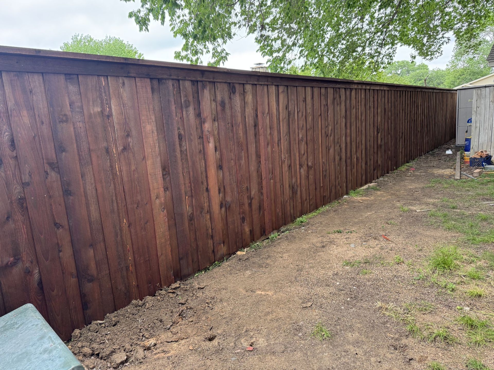Brown wooden fence in backyard, separating dirt path and grass, under a tree.