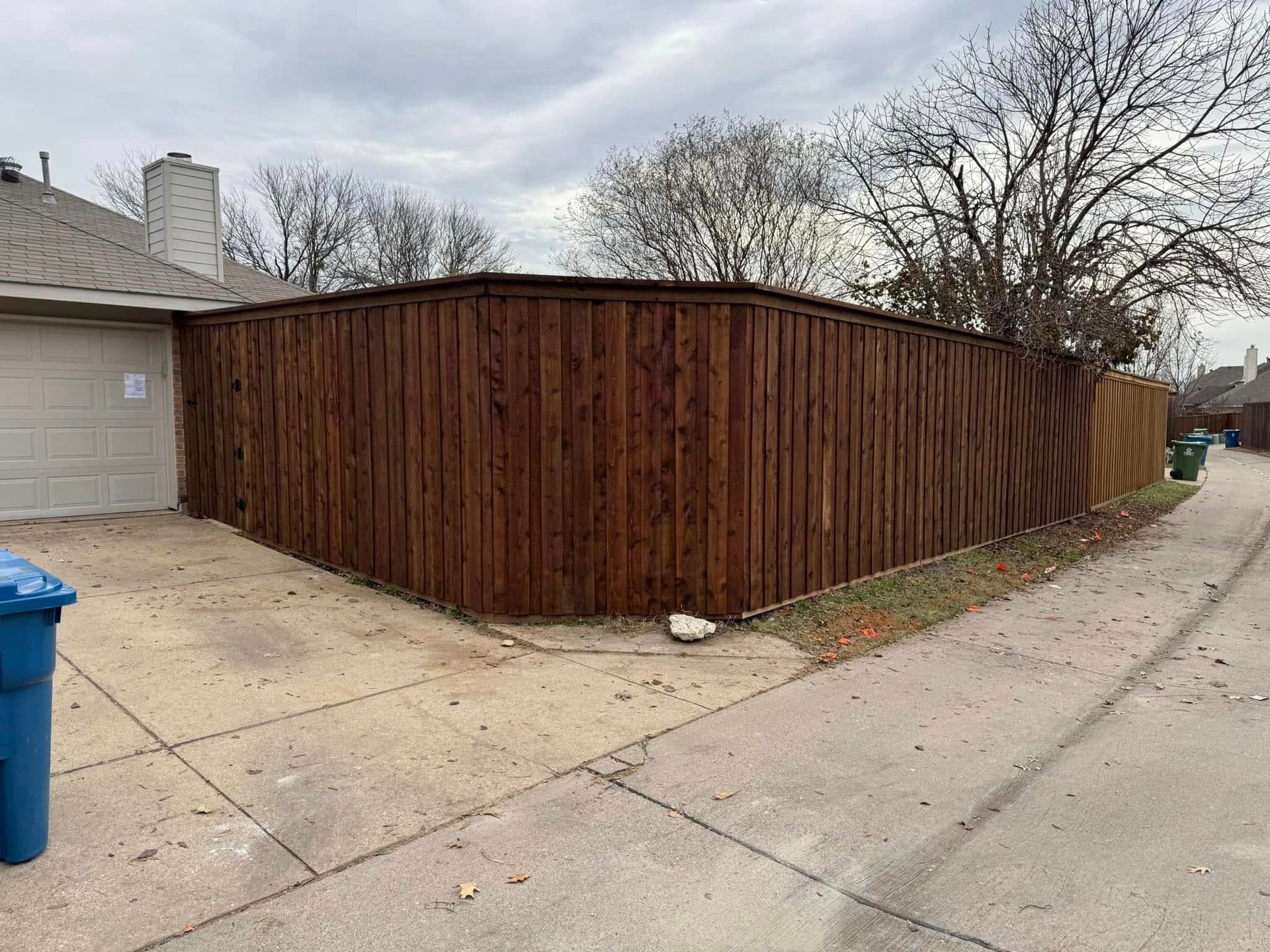 Dark brown wooden fence, cornering driveway, cloudy sky, trash bins.