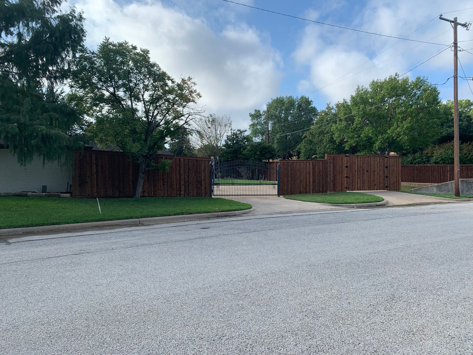 Brown wooden fence with a black gated entrance on a street with green grass and trees. Cloudy sky.