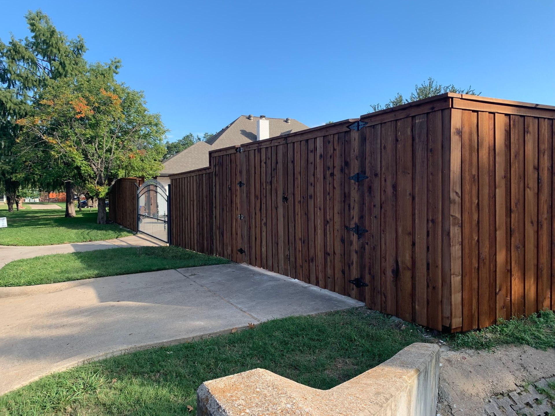 Brown wooden fence beside concrete driveway and green lawn under a blue sky.