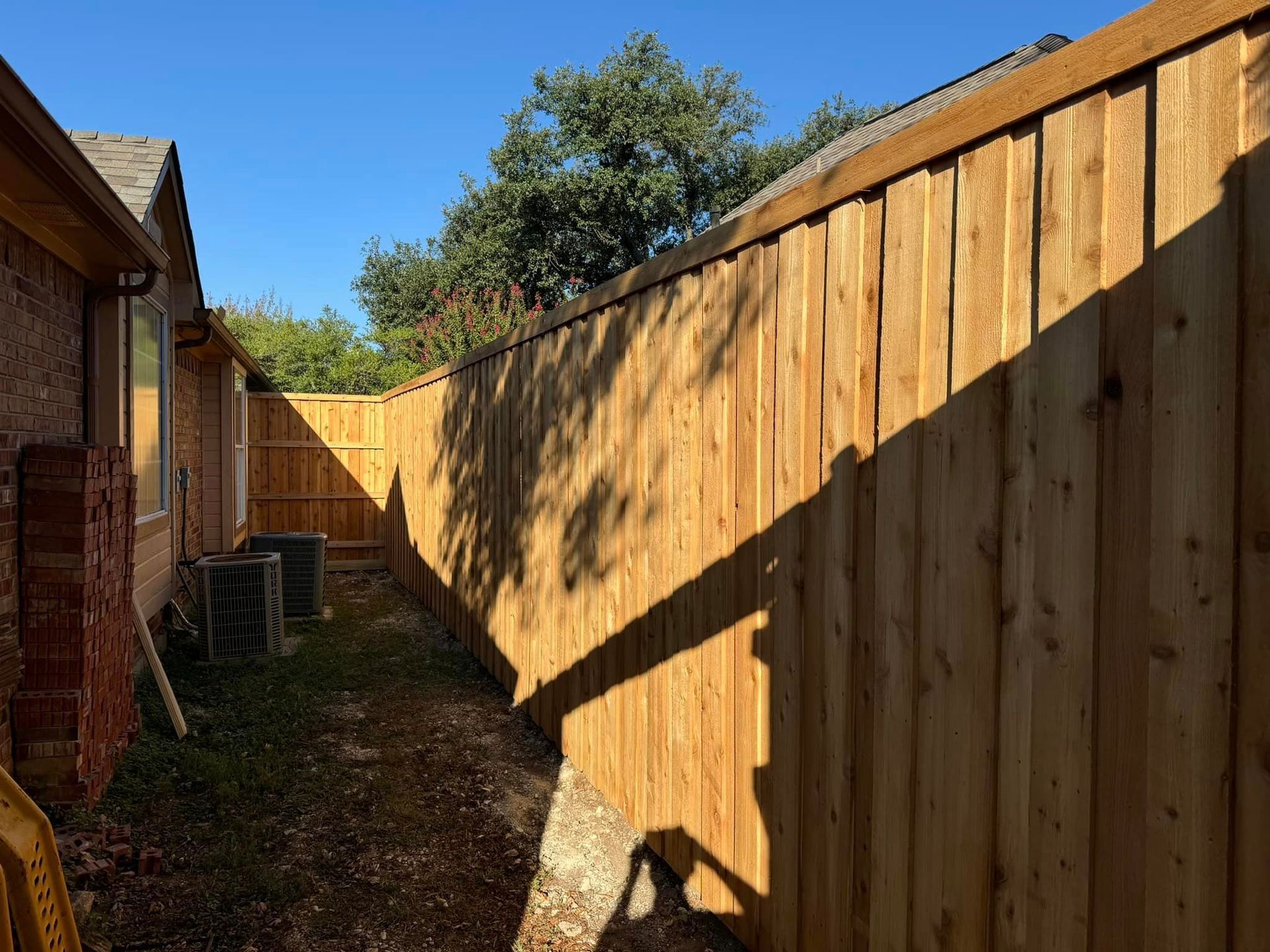 Wooden fence next to a brick house. Bright sunlight casts long shadows.