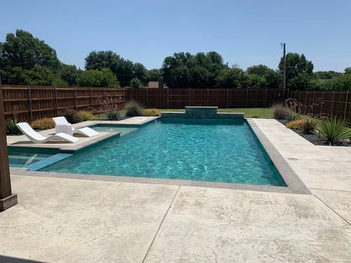 Backyard pool with white lounge chairs, concrete patio, and brown wooden fence.