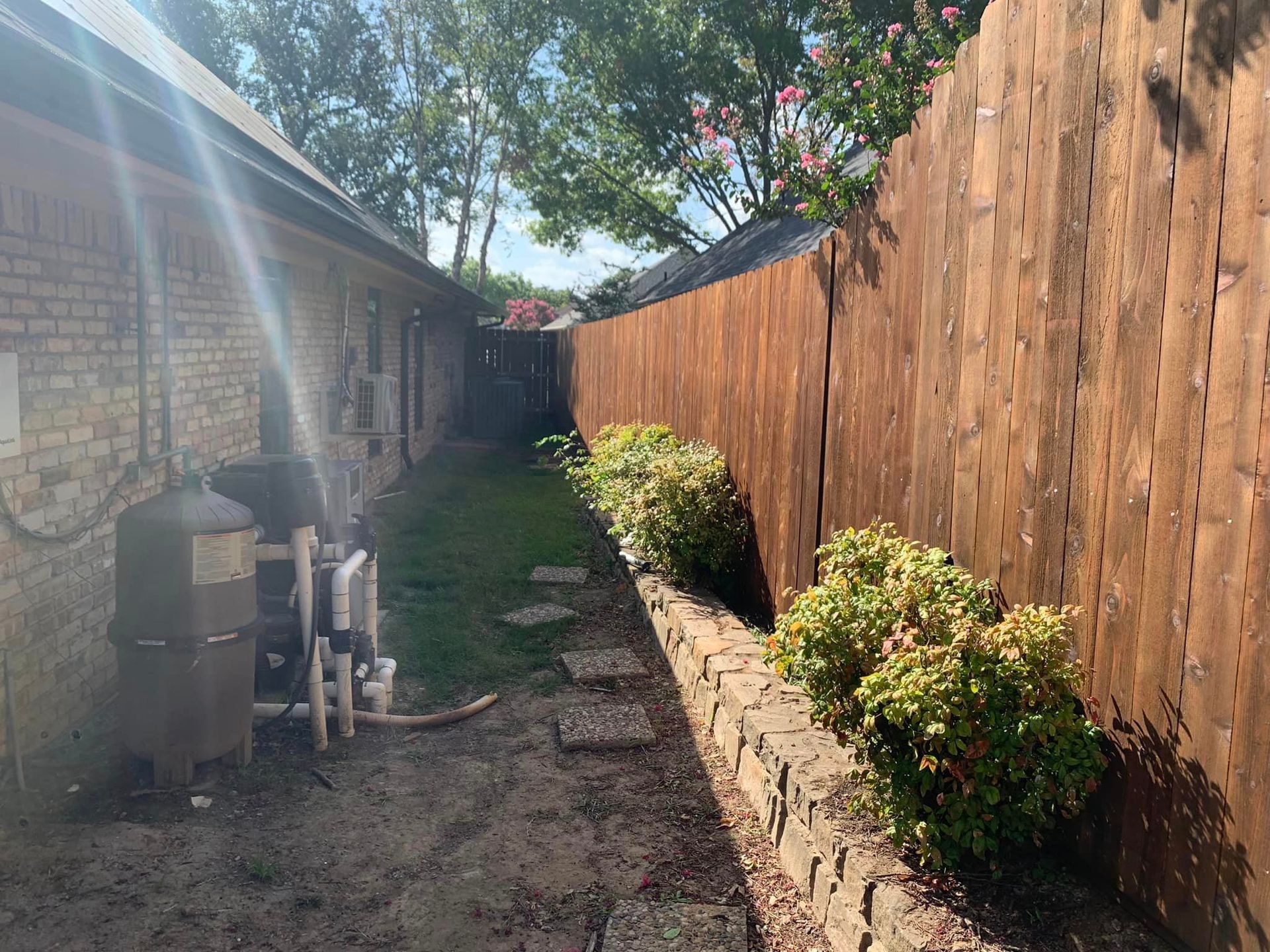 Narrow backyard with a brown fence, brick house, small stone walkway, and bushes.