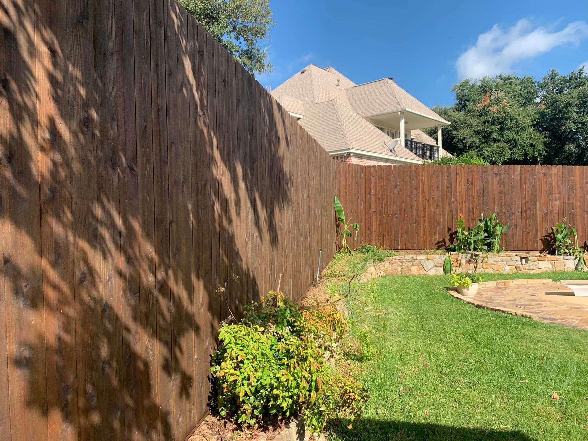 Brown wooden fence in a backyard with green grass, small plants, and a house in the background.