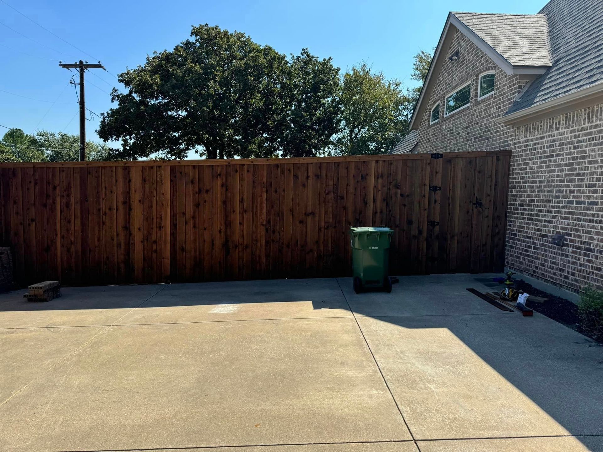 Wooden fence, green trash bin, and brick house in a sunny backyard.