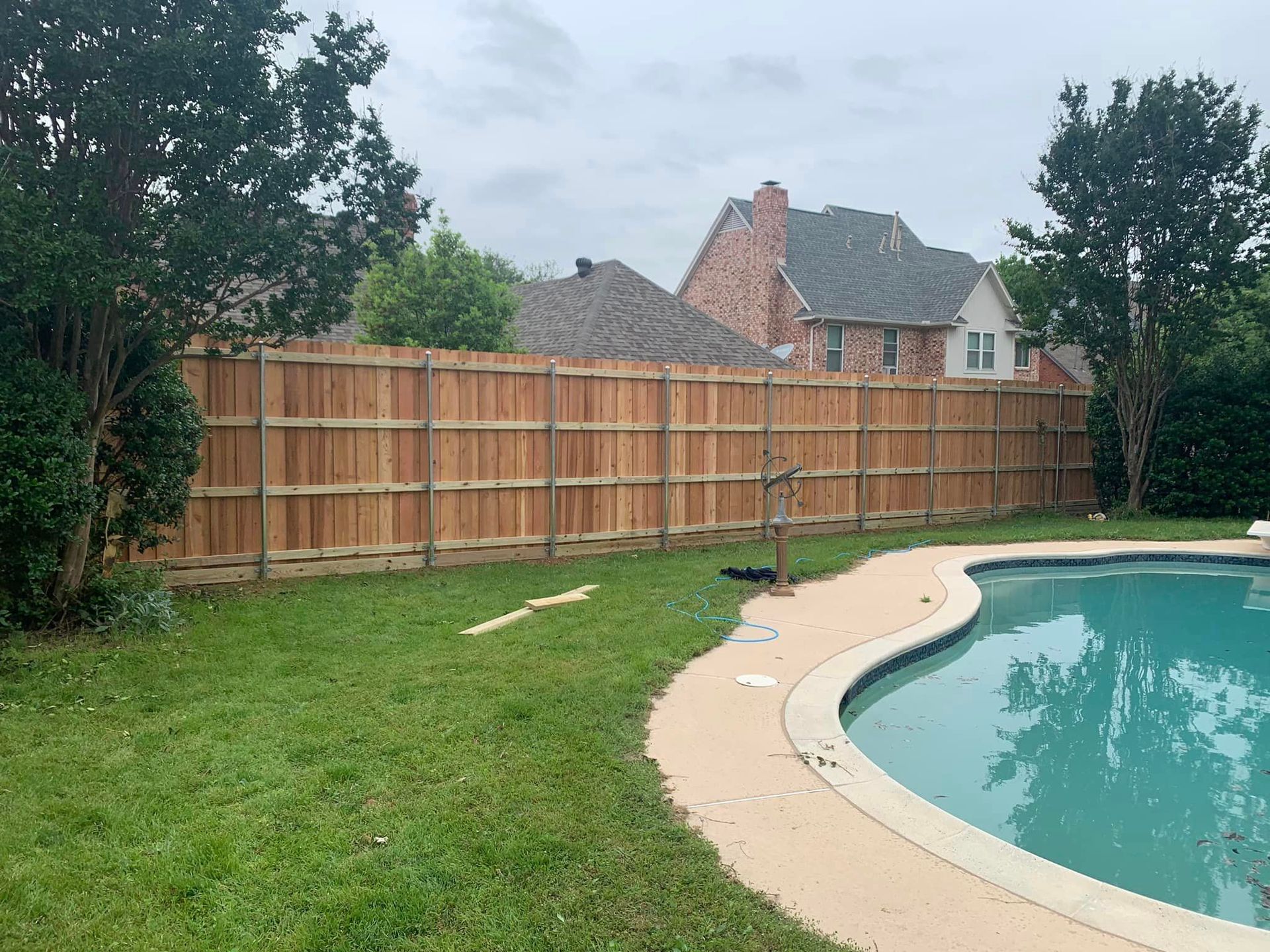 Wooden fence surrounding a backyard with a pool and lush green grass on an overcast day.