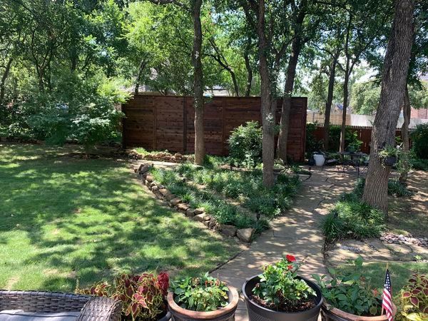 Lush backyard with green grass, trees, stone path, flower beds, and a brick fence.