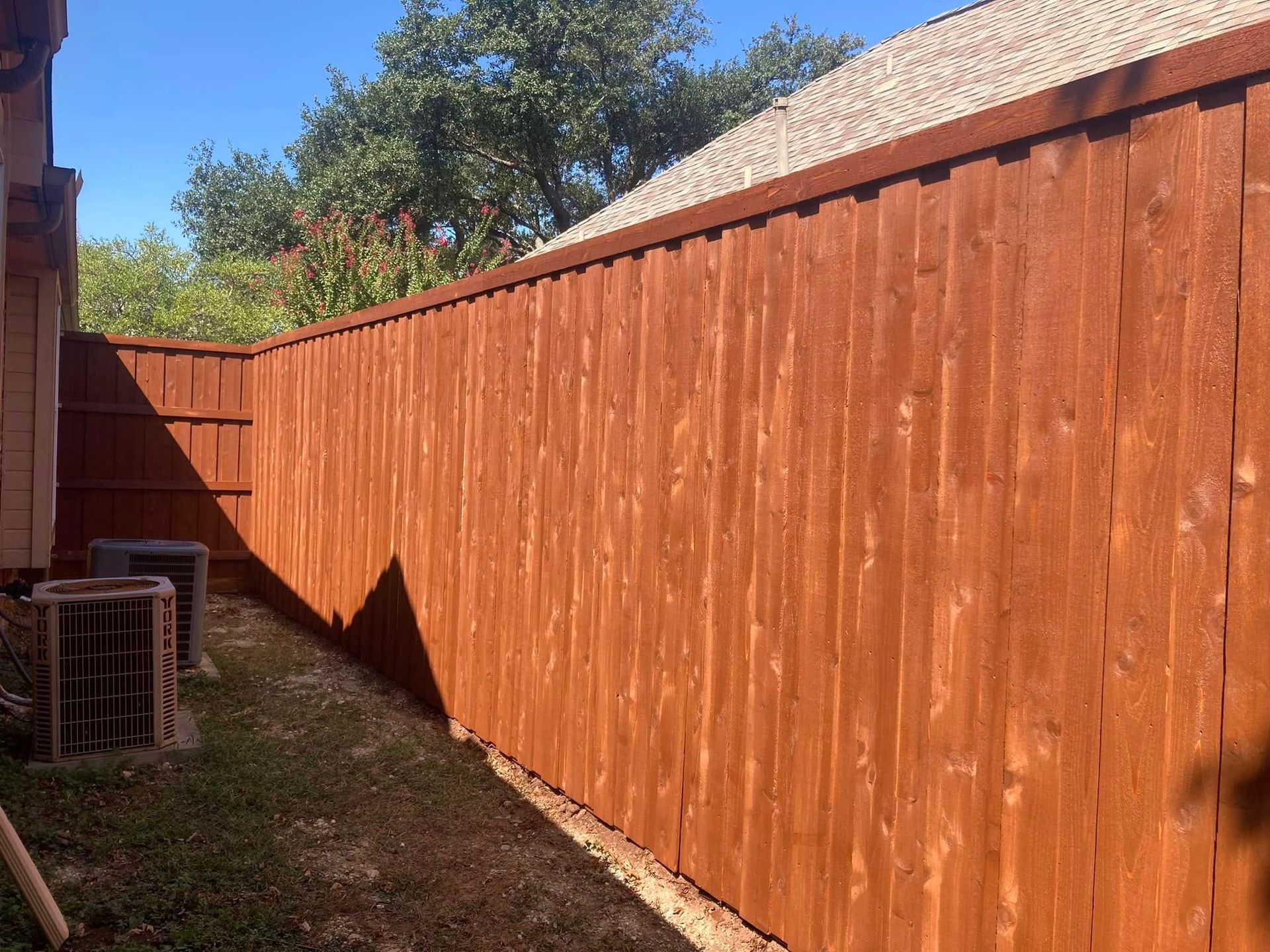 A freshly stained brown wooden fence next to a building and air conditioning units, with grass in the foreground.