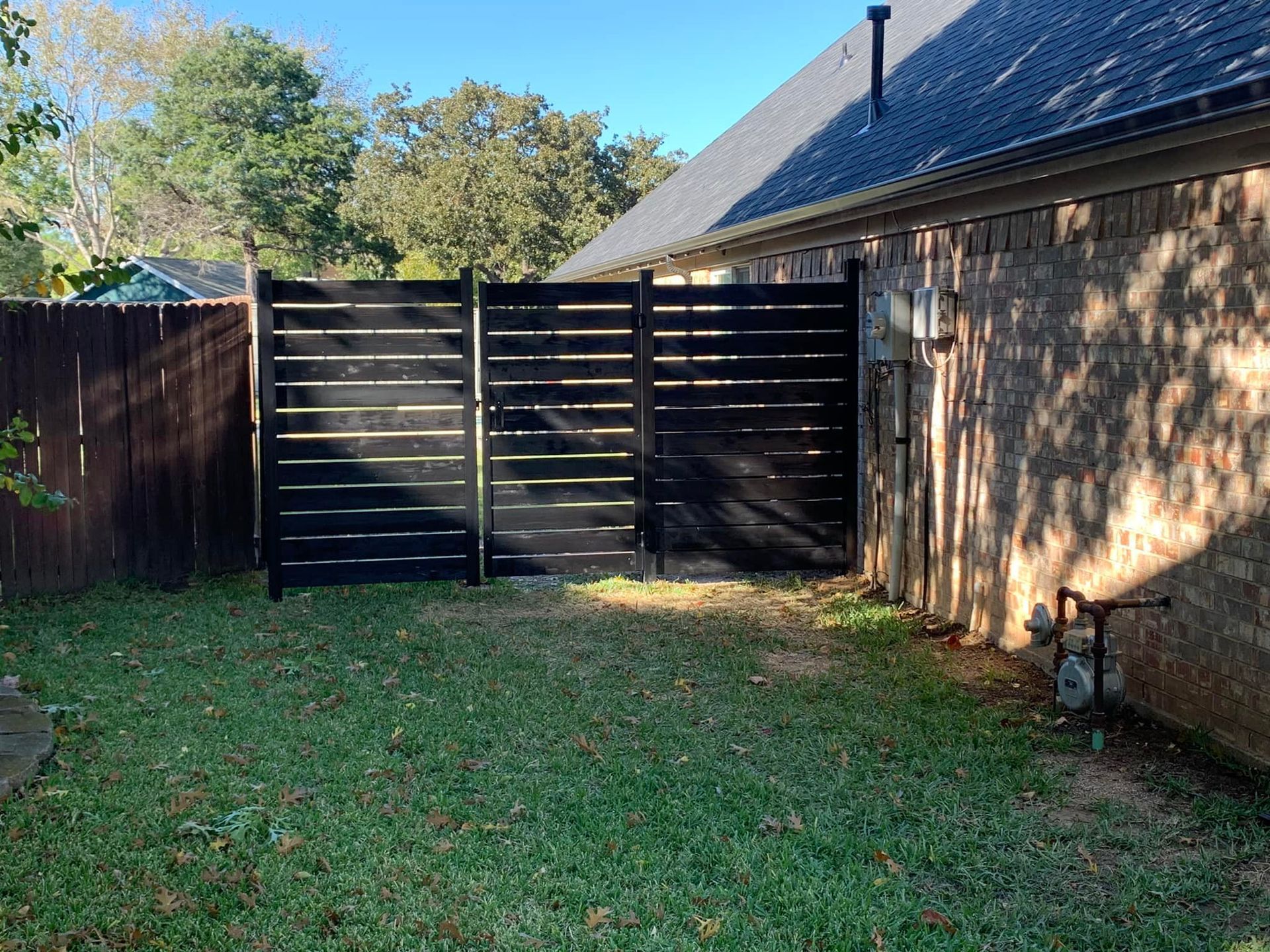 Black horizontal slat fence in backyard against brick wall and brown fence.