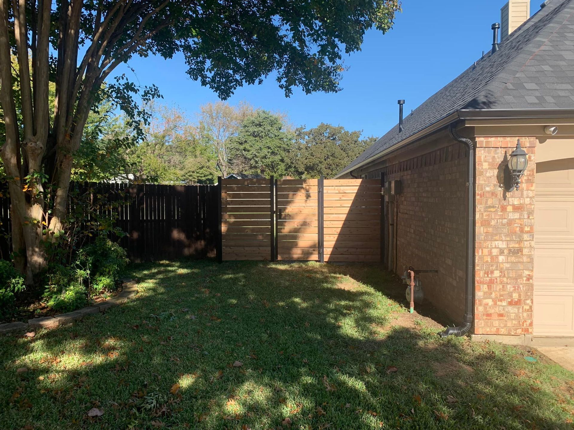 Backyard with a fence, grass, a brick house, and a tree on a sunny day.