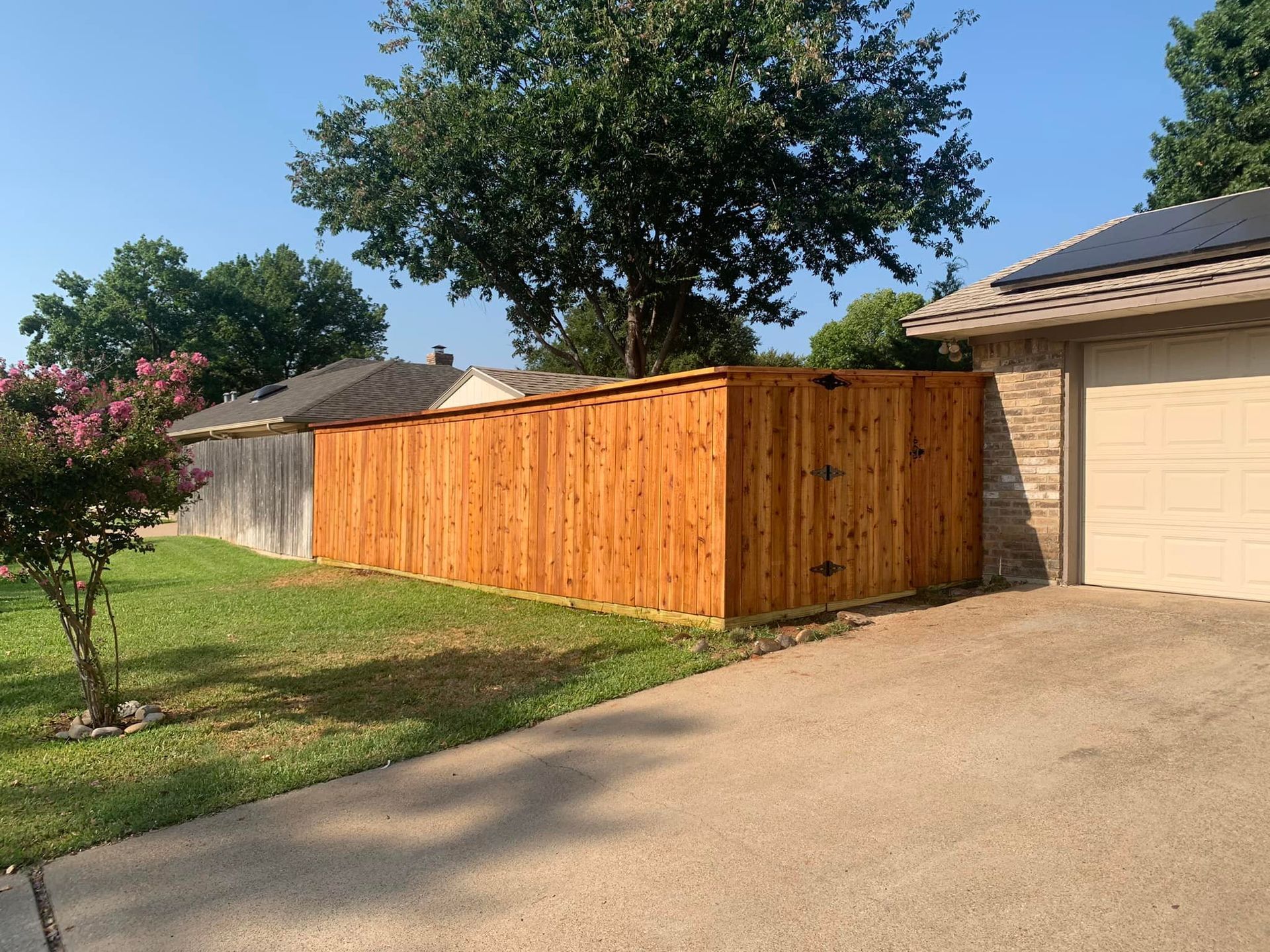 Wooden fence next to a driveway, with a lighter color than the neighboring weathered fence, under a sunny sky.