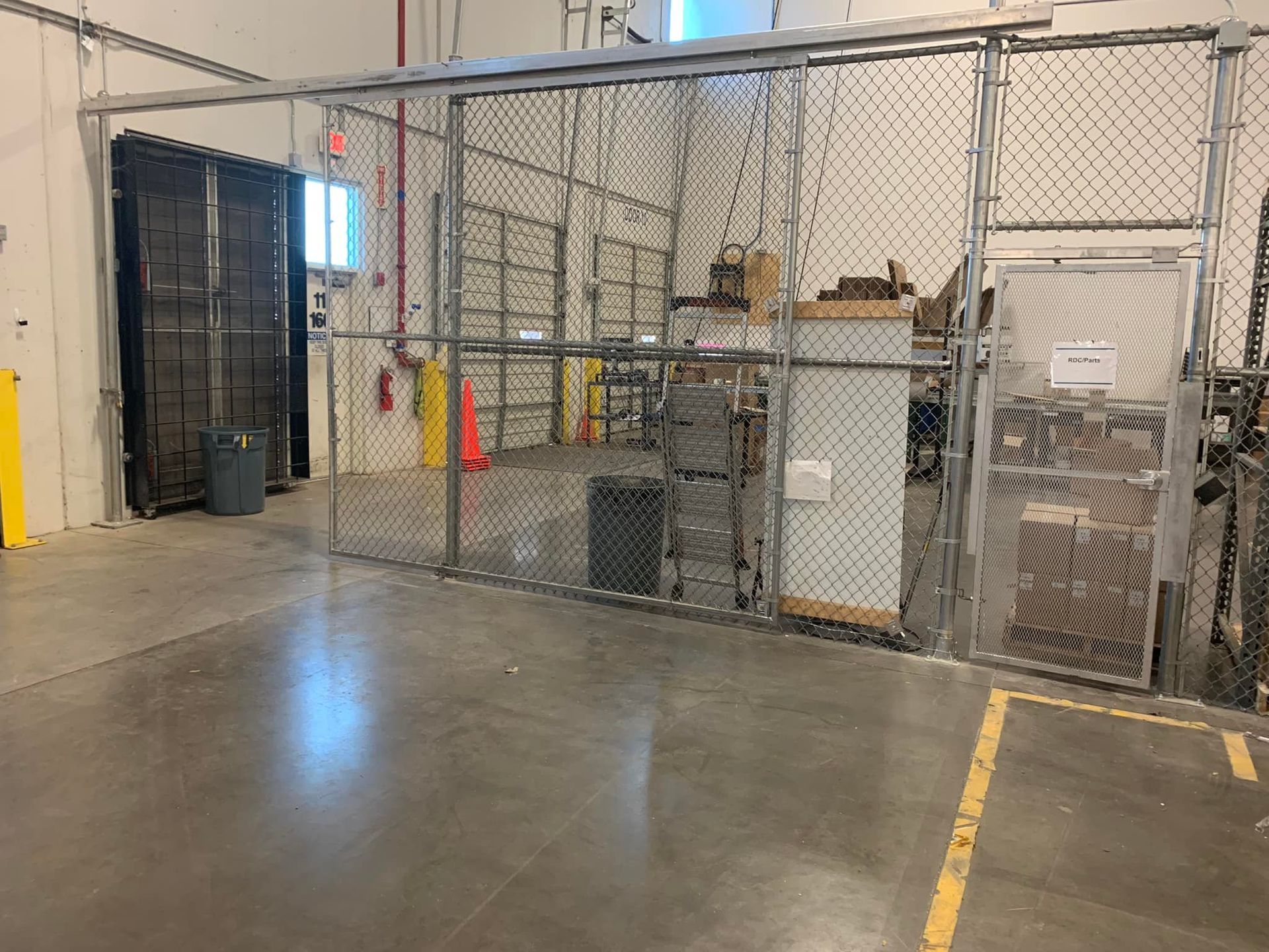 A chain-link fenced-in area in a warehouse. Inside, a gray cabinet, boxes, and a trash can are visible.