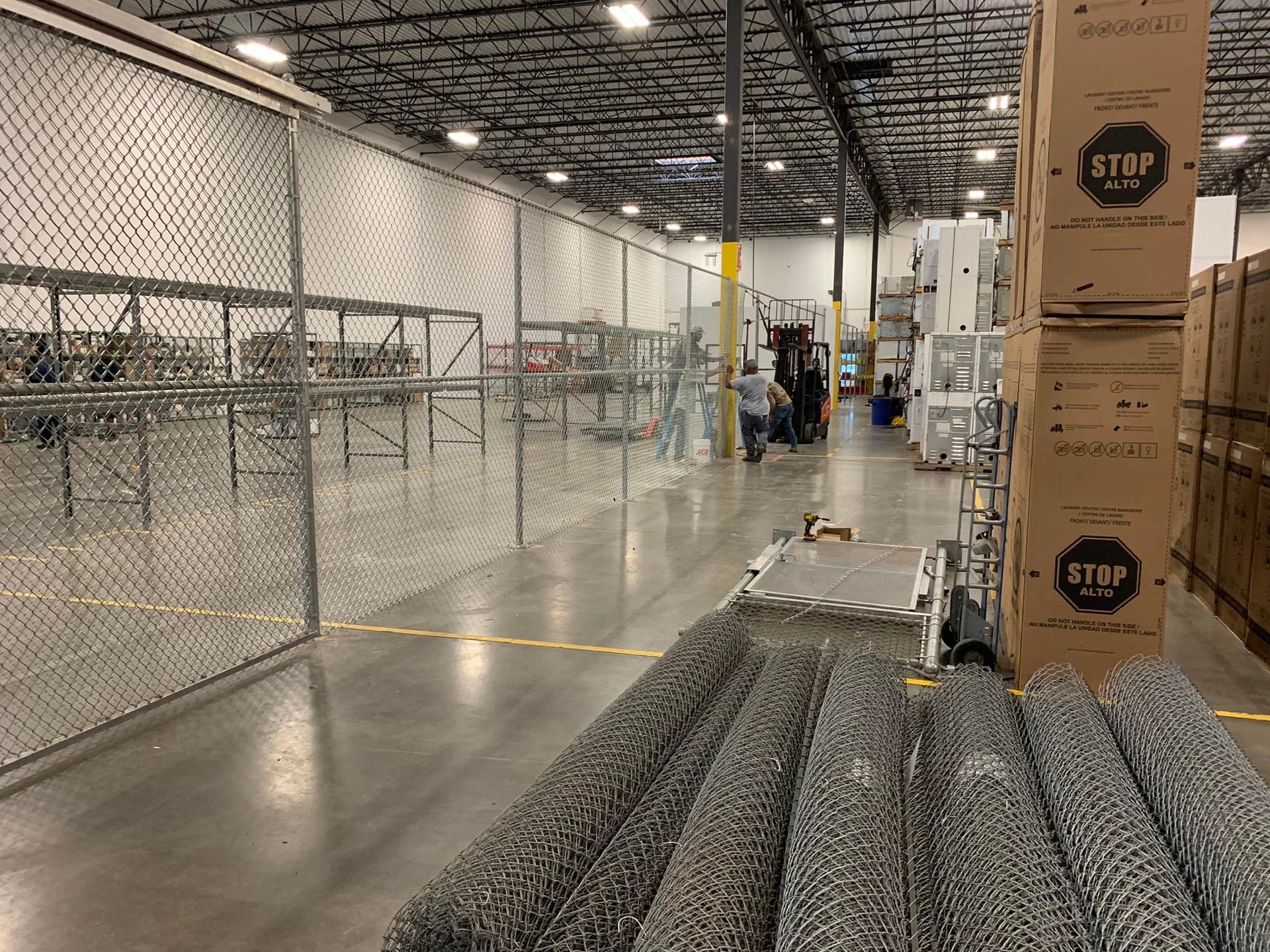Warehouse interior with chain link fencing, workers, stacked boxes, and rolls of wire mesh.