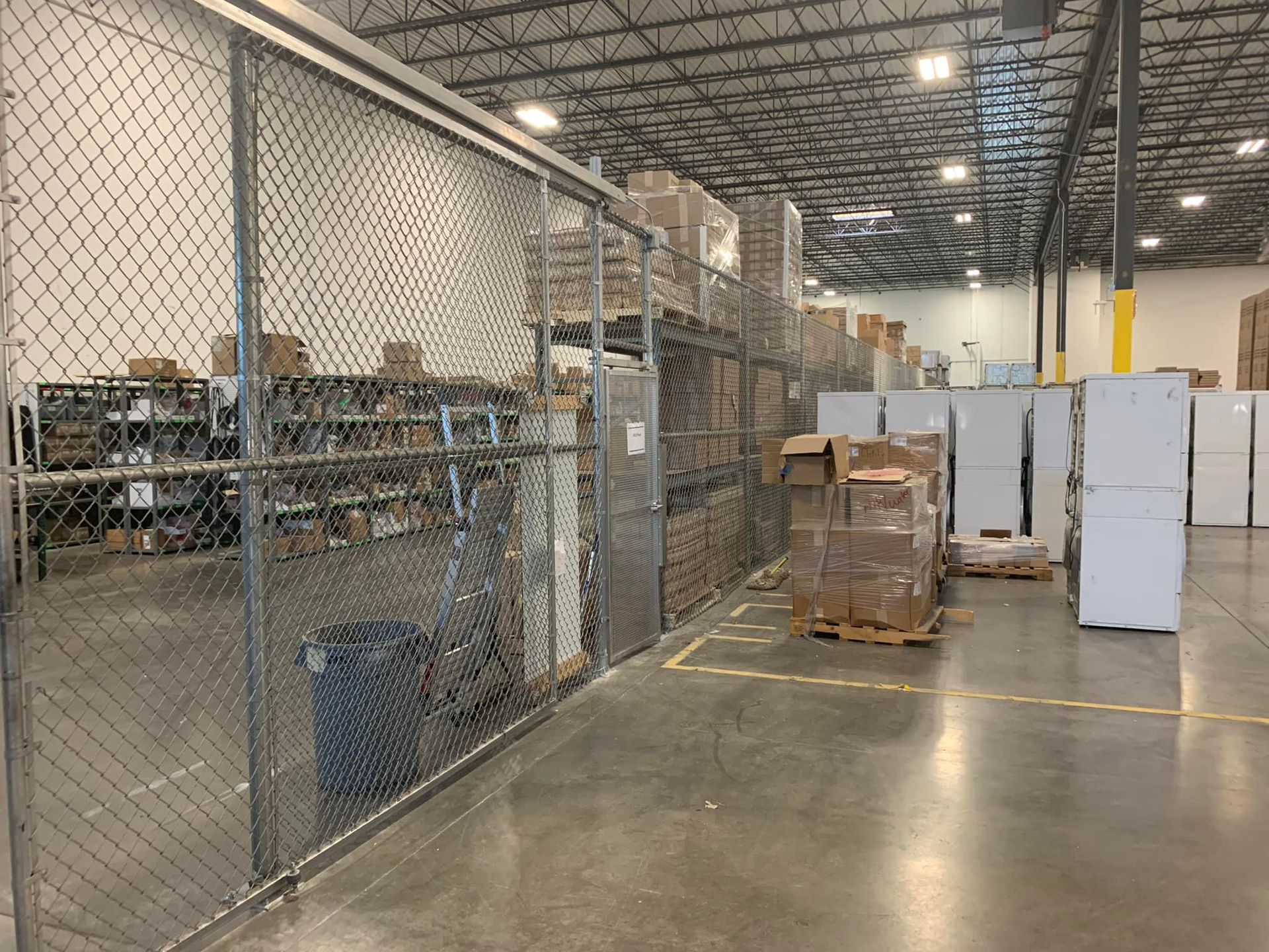 Warehouse interior; chain-link fence encloses shelving with boxes. Concrete floor, boxes on pallets, white appliances.
