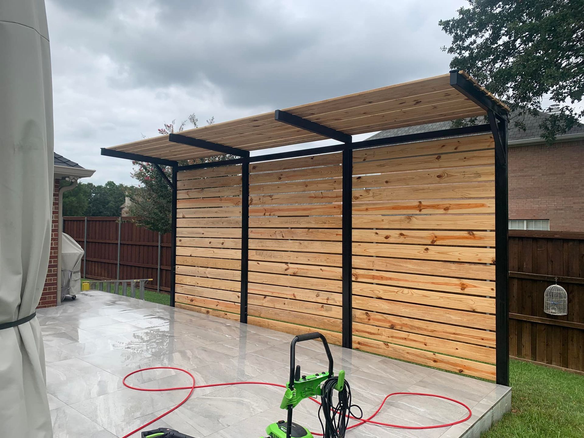 Wooden pergola with horizontal planks, black metal frame, on a gray patio with a pressure washer.