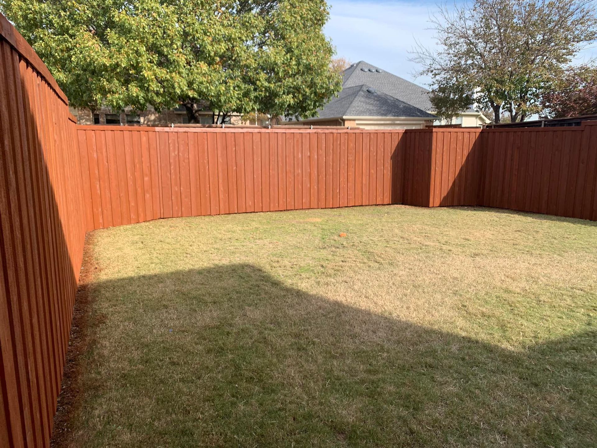A fenced backyard with brown wooden fence, green and brown grass, and a house in the background.