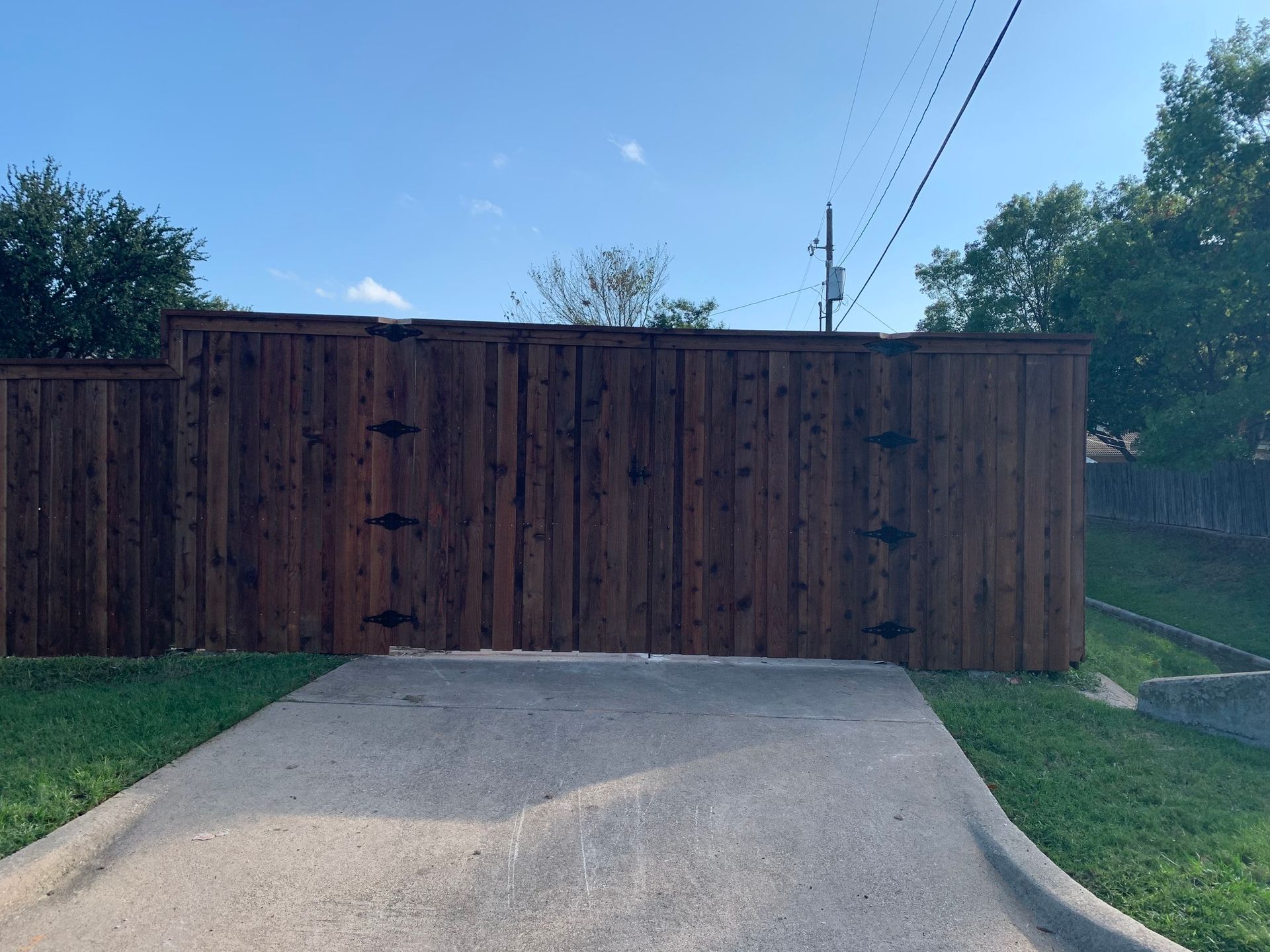 A dark brown wooden fence with decorative hinges stands in front of a driveway, blue sky overhead.