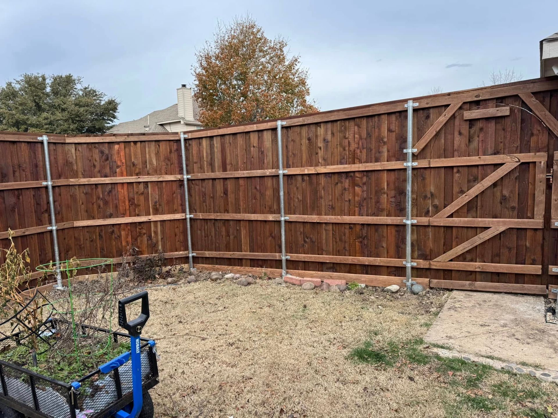 A wooden fence with a gate in a backyard, with metal supports and a brown stain.