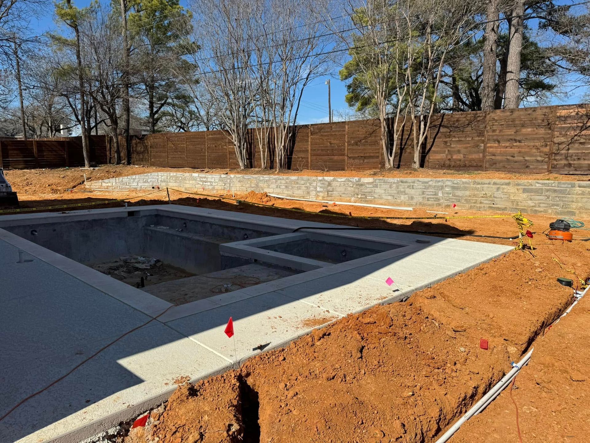 Pool under construction with concrete edges, dirt, and a wooden fence in the background.