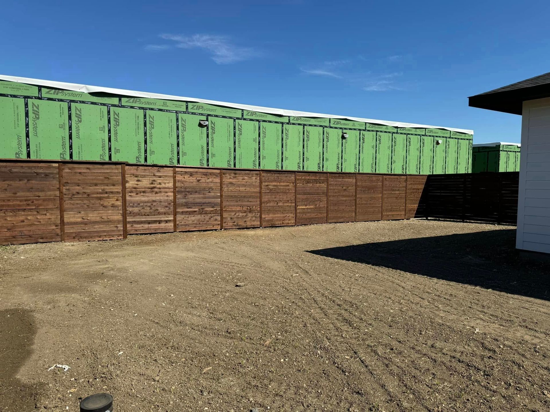 Brown wooden fence in front of green-sheathed construction, set on gravel against a blue sky.