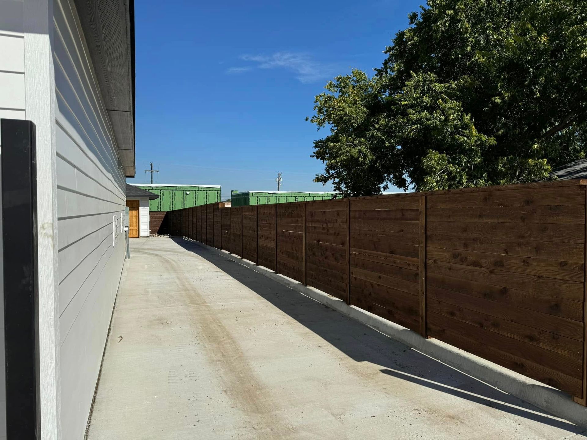 Concrete driveway beside a light-colored building and a stained wooden fence under a blue sky.