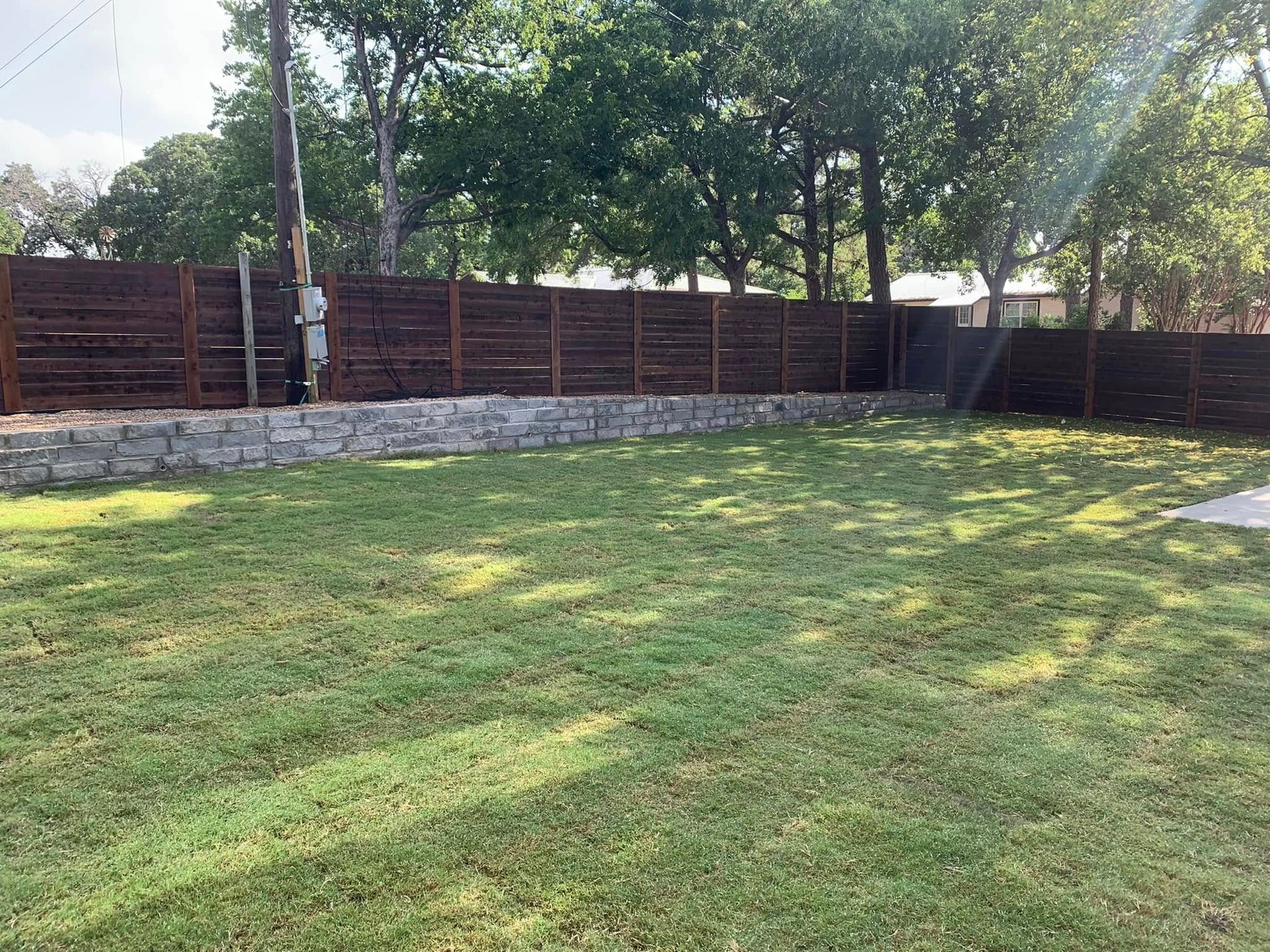 Lawn with a dark wooden fence on top of a stone retaining wall. Trees in background.