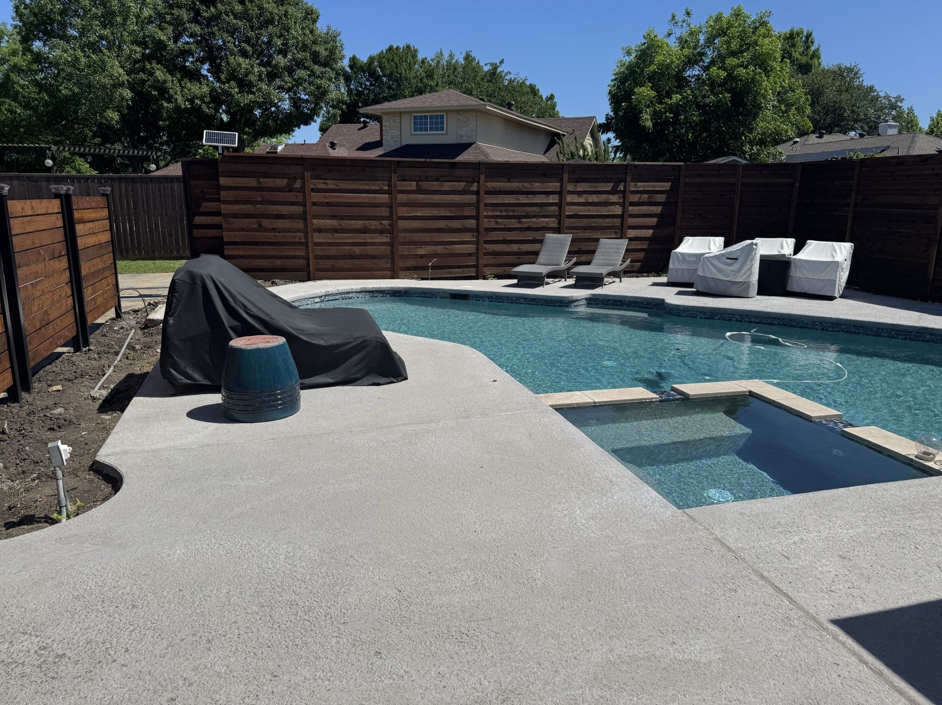 Pool area with patio furniture, pool, and hot tub, surrounded by a wooden fence and trees under a blue sky.