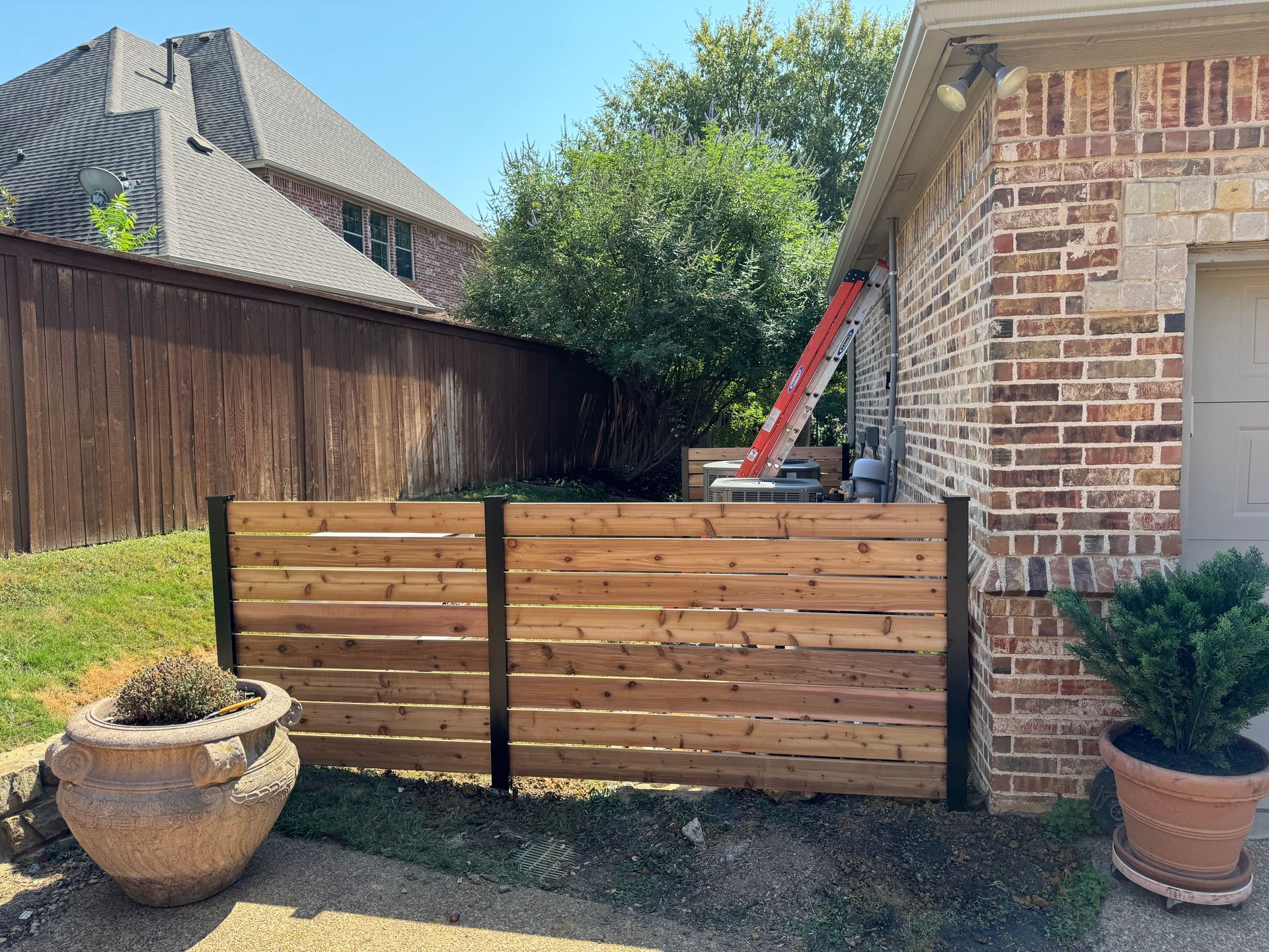 Wooden horizontal slat fence, brick house, ladder propped against the wall, potted plants.