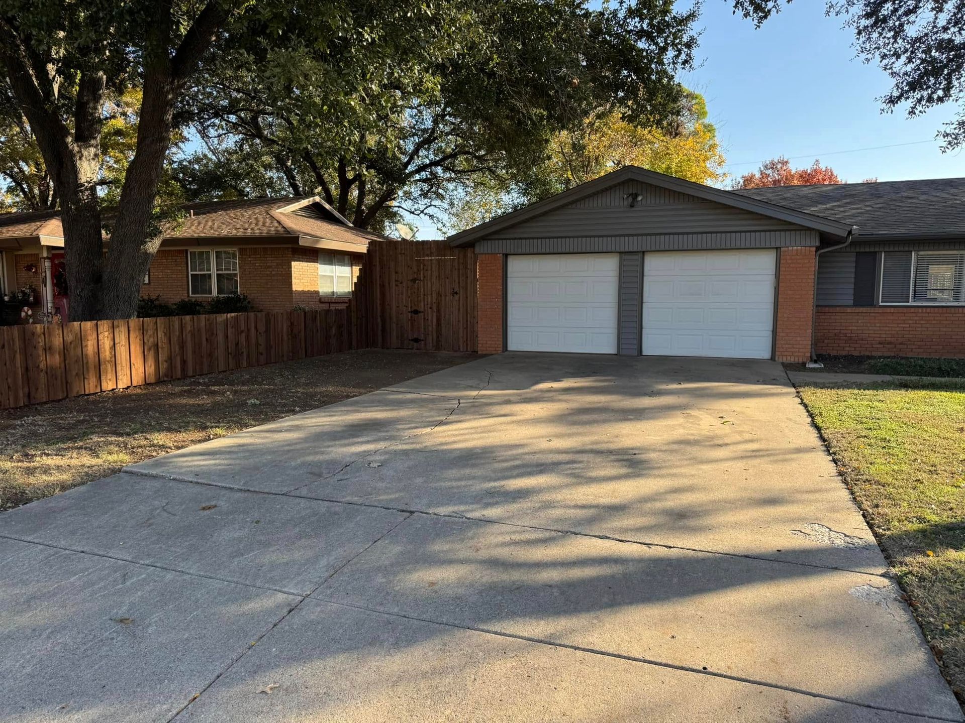 A house with a two-car garage and driveway on a sunny day. A wooden fence and trees are also visible.