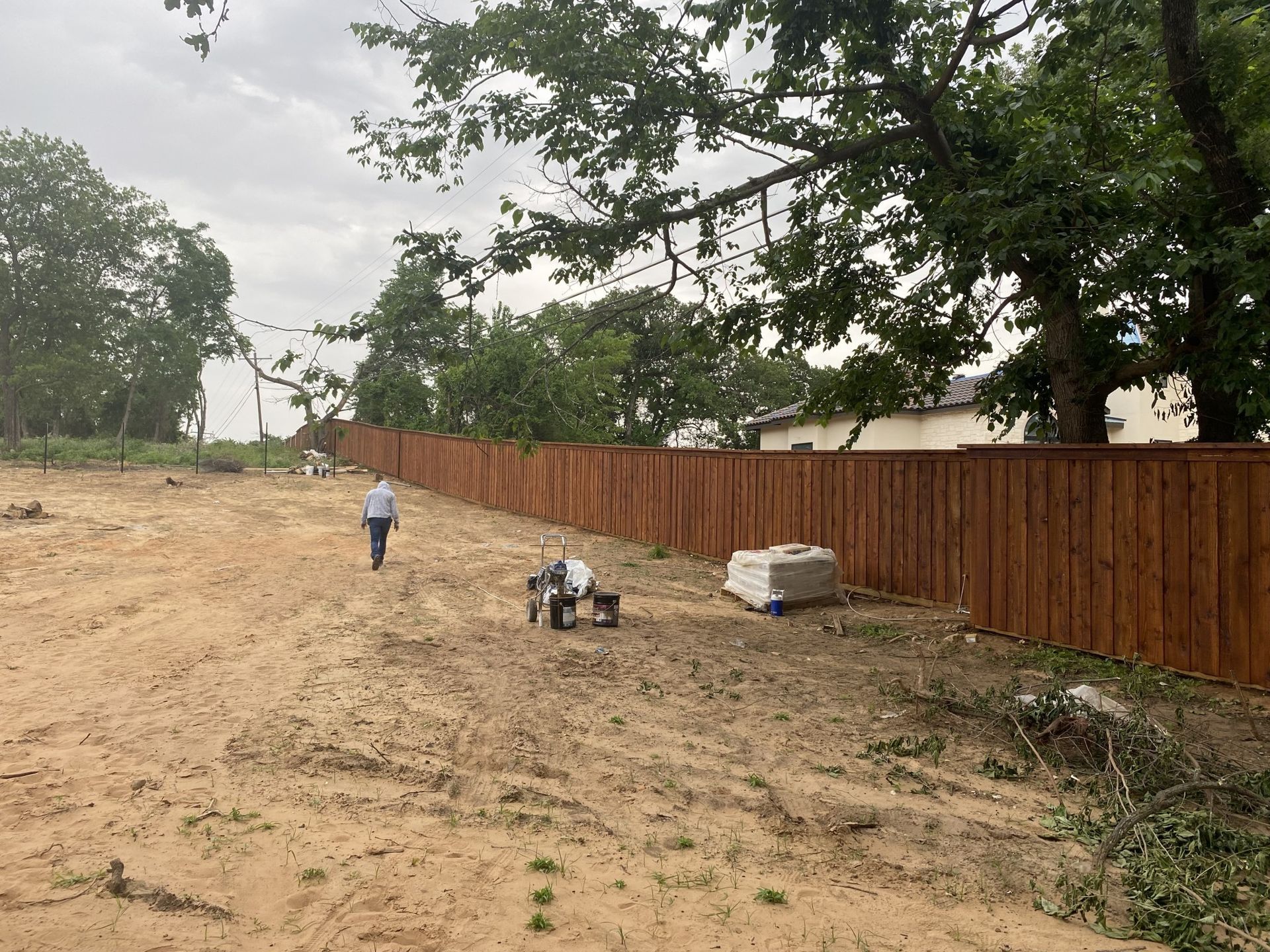 Person walking on dirt path near a brown wooden fence with a small machine and concrete blocks. Cloudy sky.