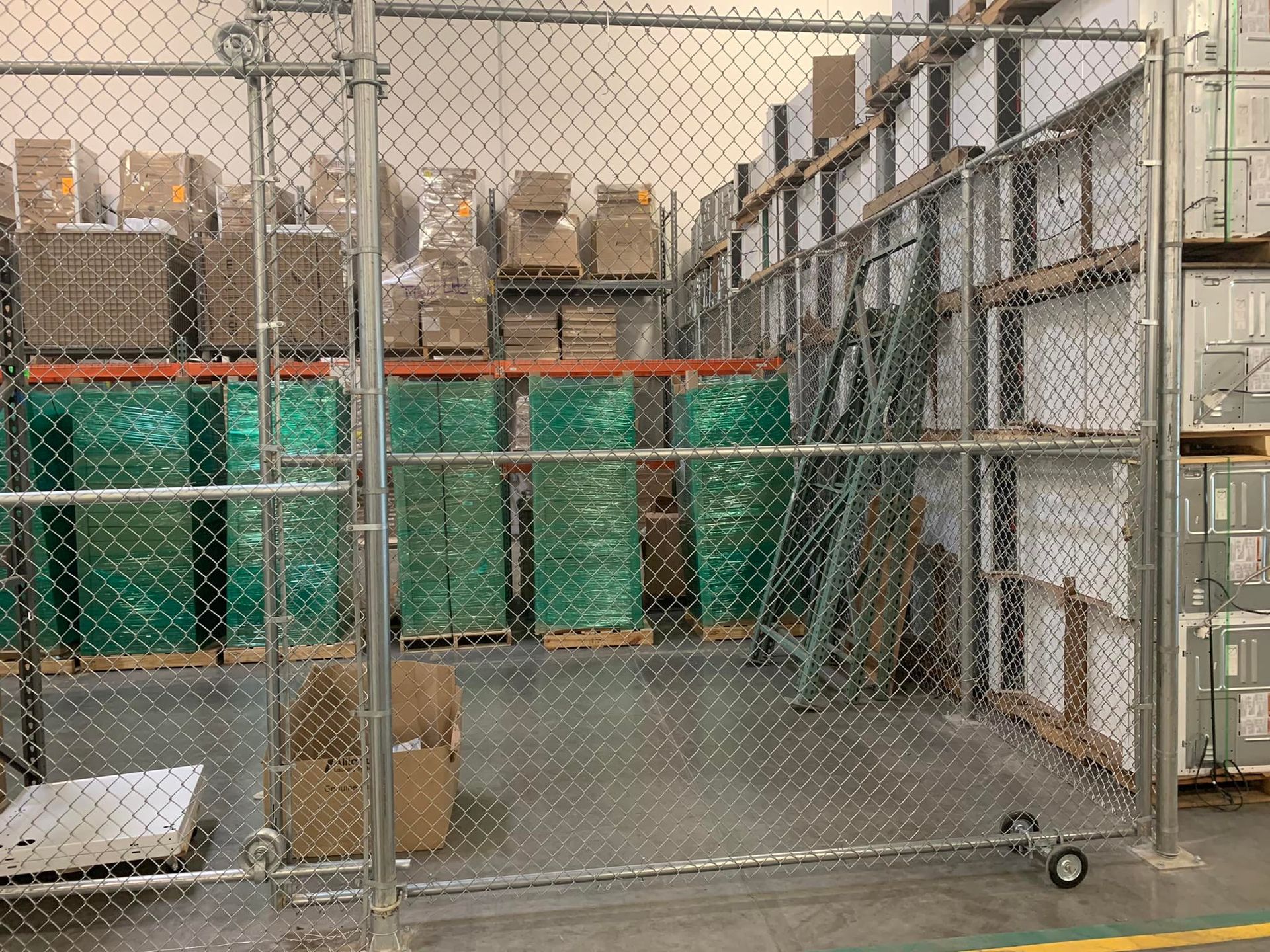 Chain-link fence encloses pallets of green-wrapped items in a warehouse. Boxes and shelves are visible behind the fence.