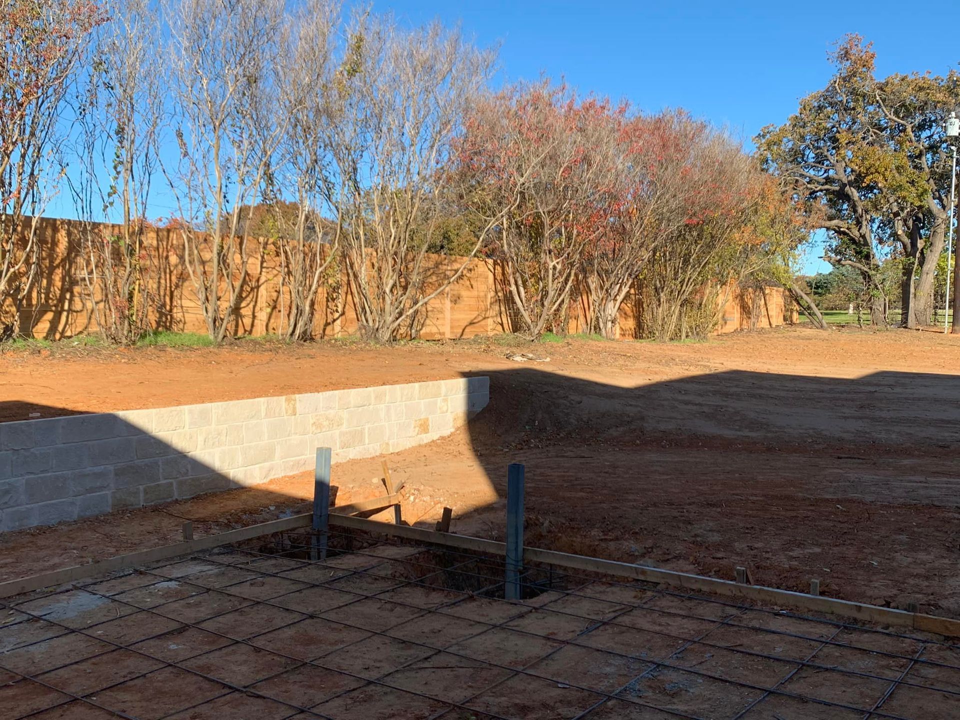 Construction site: exposed rebar grid and pipes, brick wall in background, red dirt, trees and clear sky.