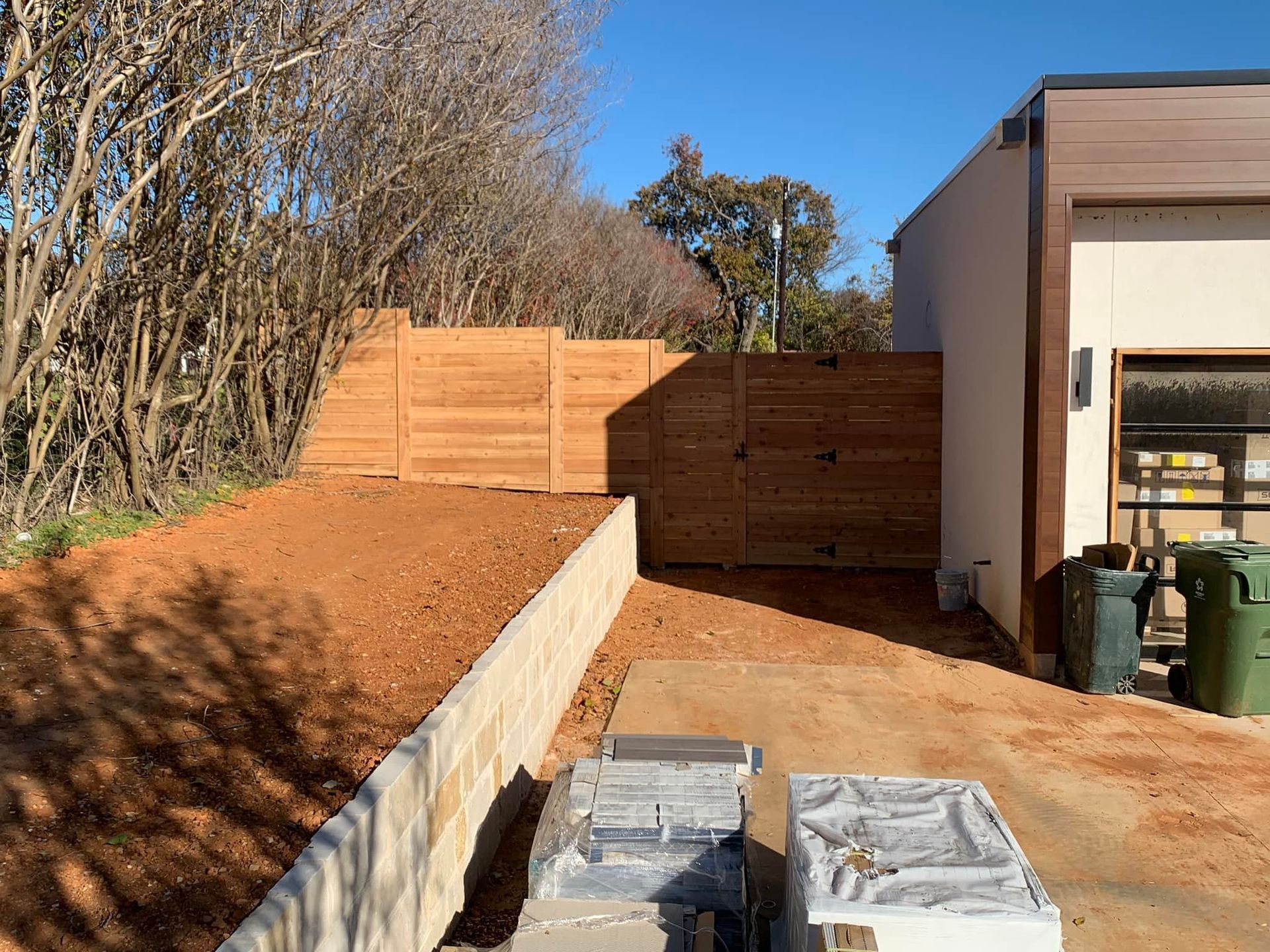 A red dirt driveway next to a cream-colored building with a wooden fence and two green trash cans.