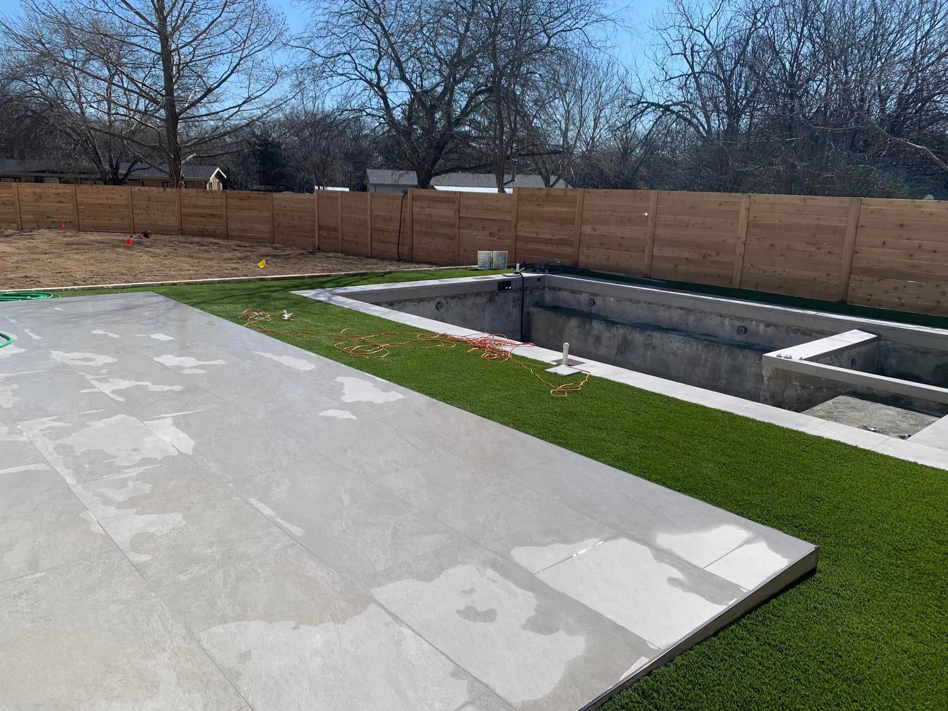 Concrete patio and pool under construction next to green artificial grass with a wooden fence in the background.