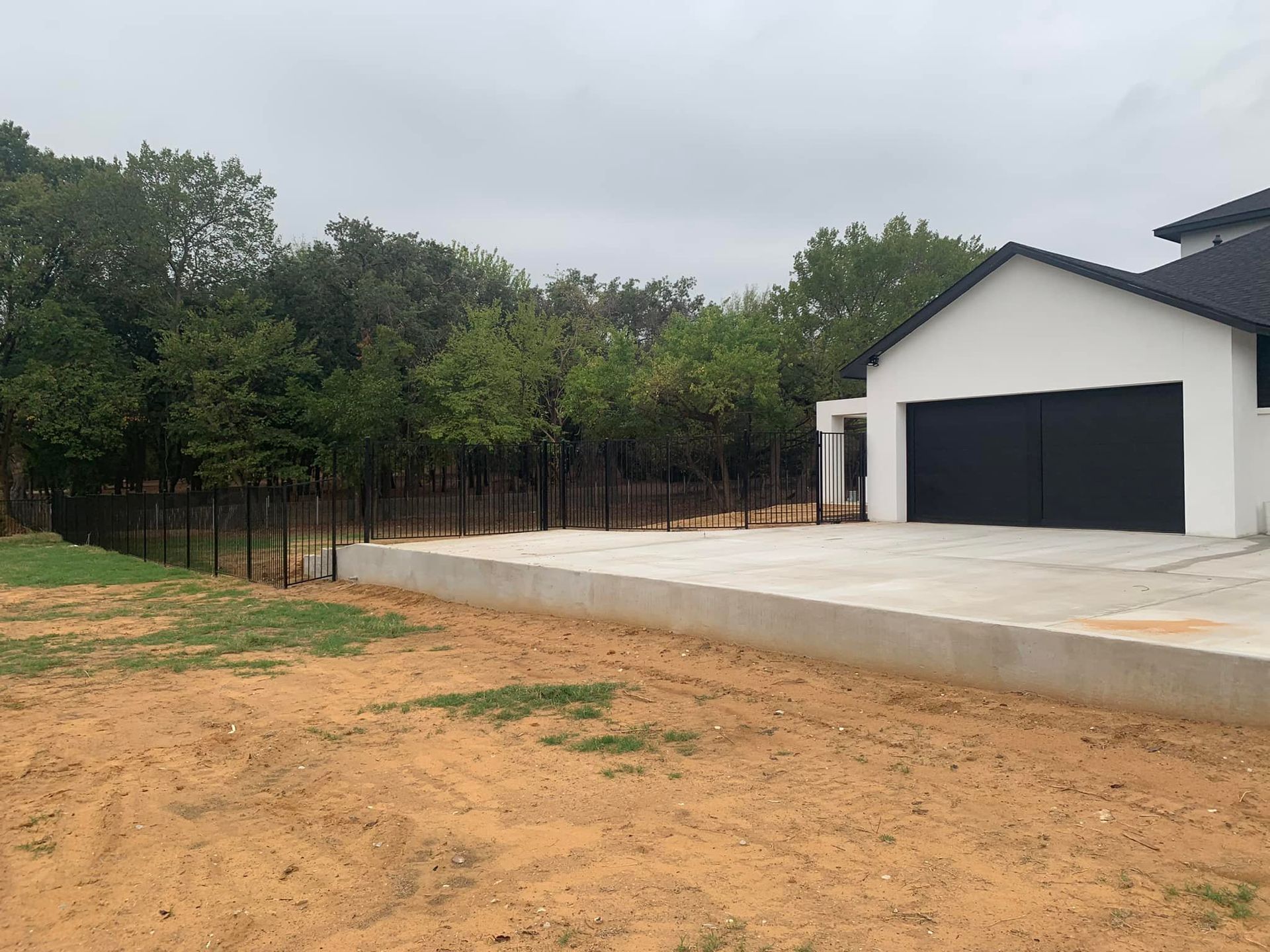 Modern white house with black garage door, concrete driveway, and fence bordering trees under a cloudy sky.