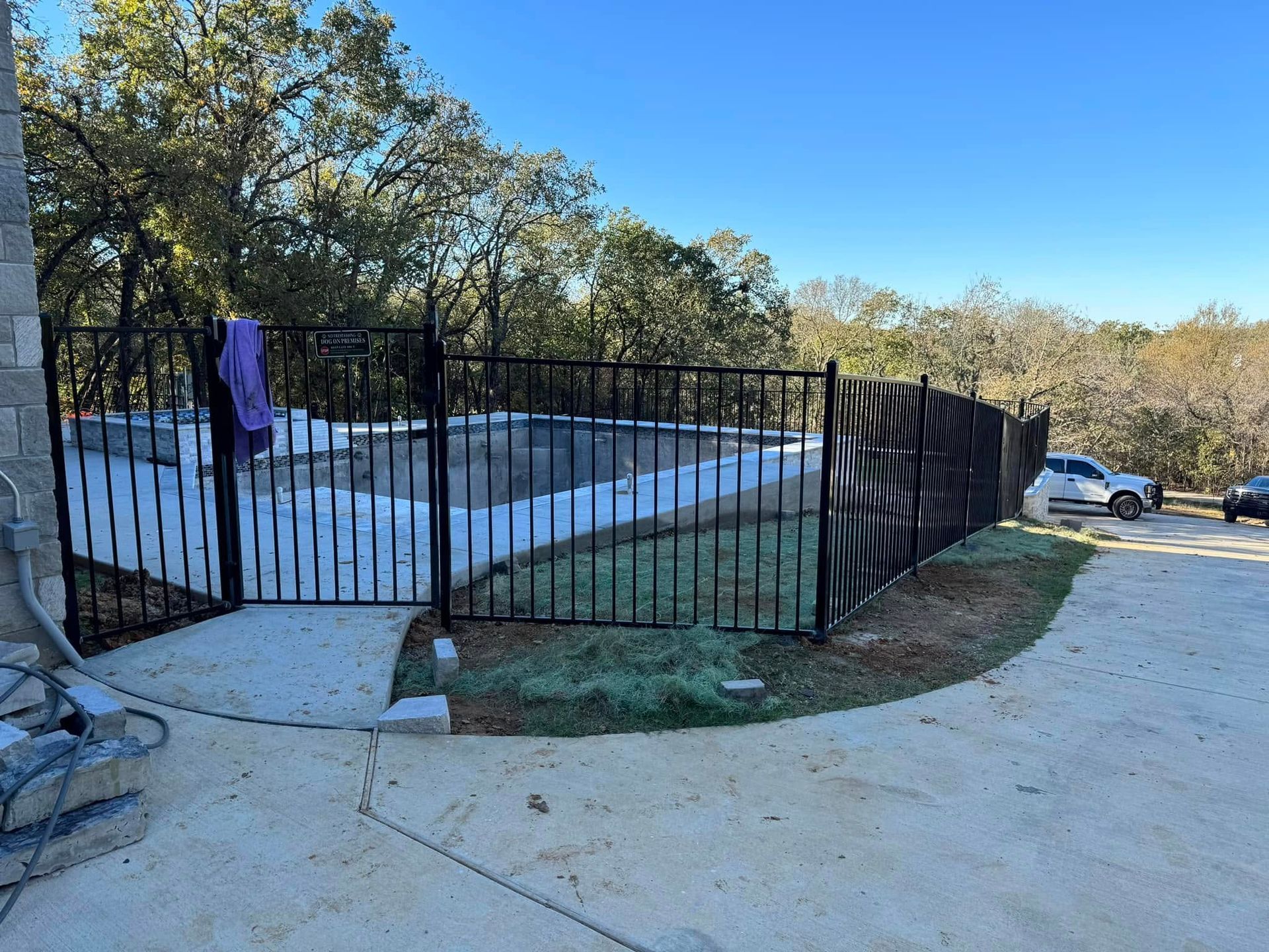 Black fence encloses a pool area with a vehicle in the background, under a clear sky.