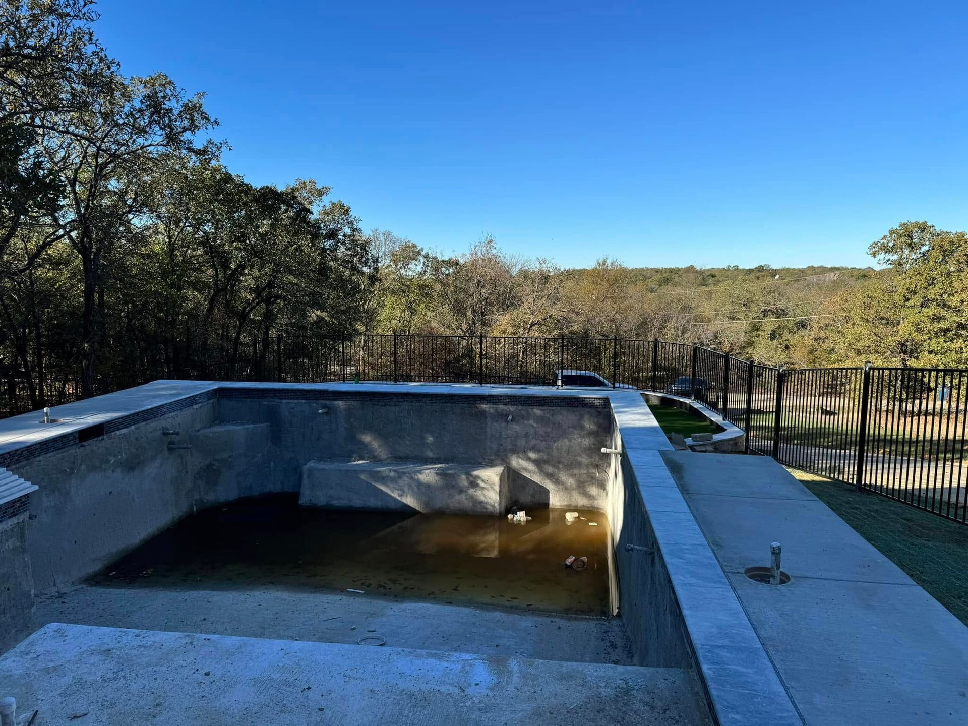 Unfinished concrete pool with dirty water, steps, and a forested landscape under a blue sky.