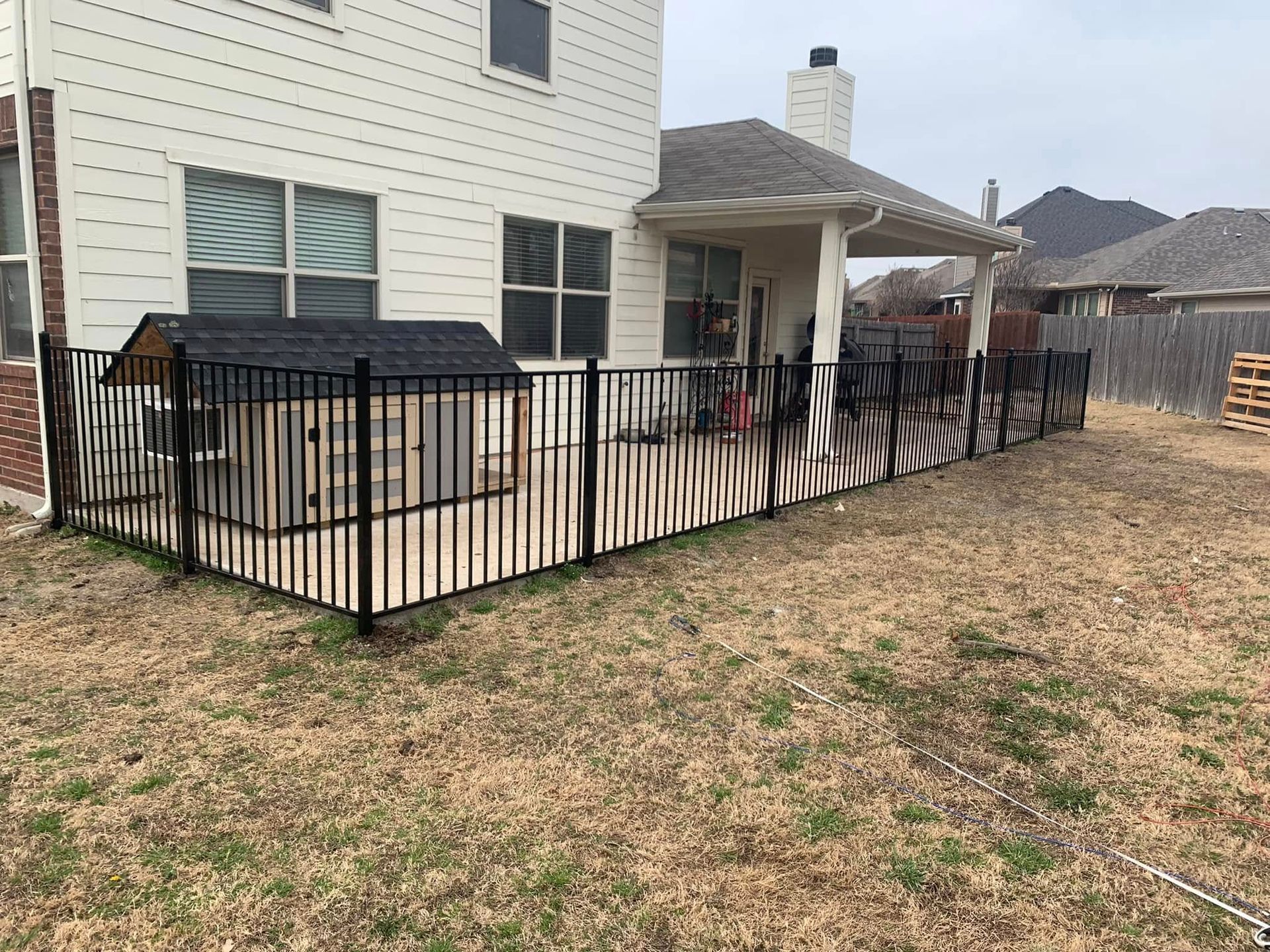 Black fenced backyard with doghouse near a house, dry grass, overcast sky.