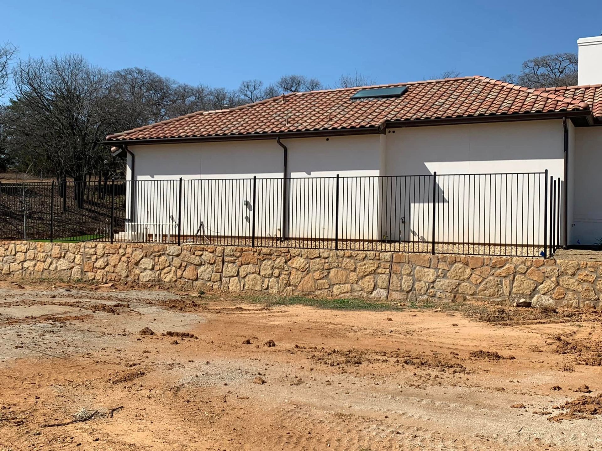 A tan house with an ornate black fence, resting on a stone wall, against a blue sky.