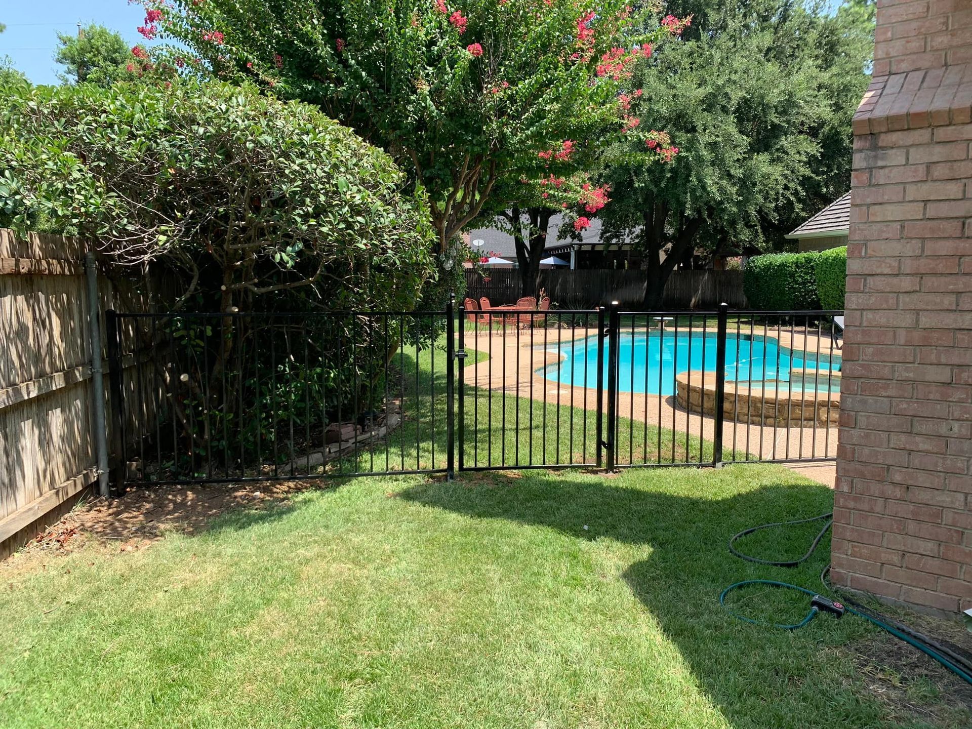 A backyard with a pool, black fence, and green grass, viewed from the side of a house.