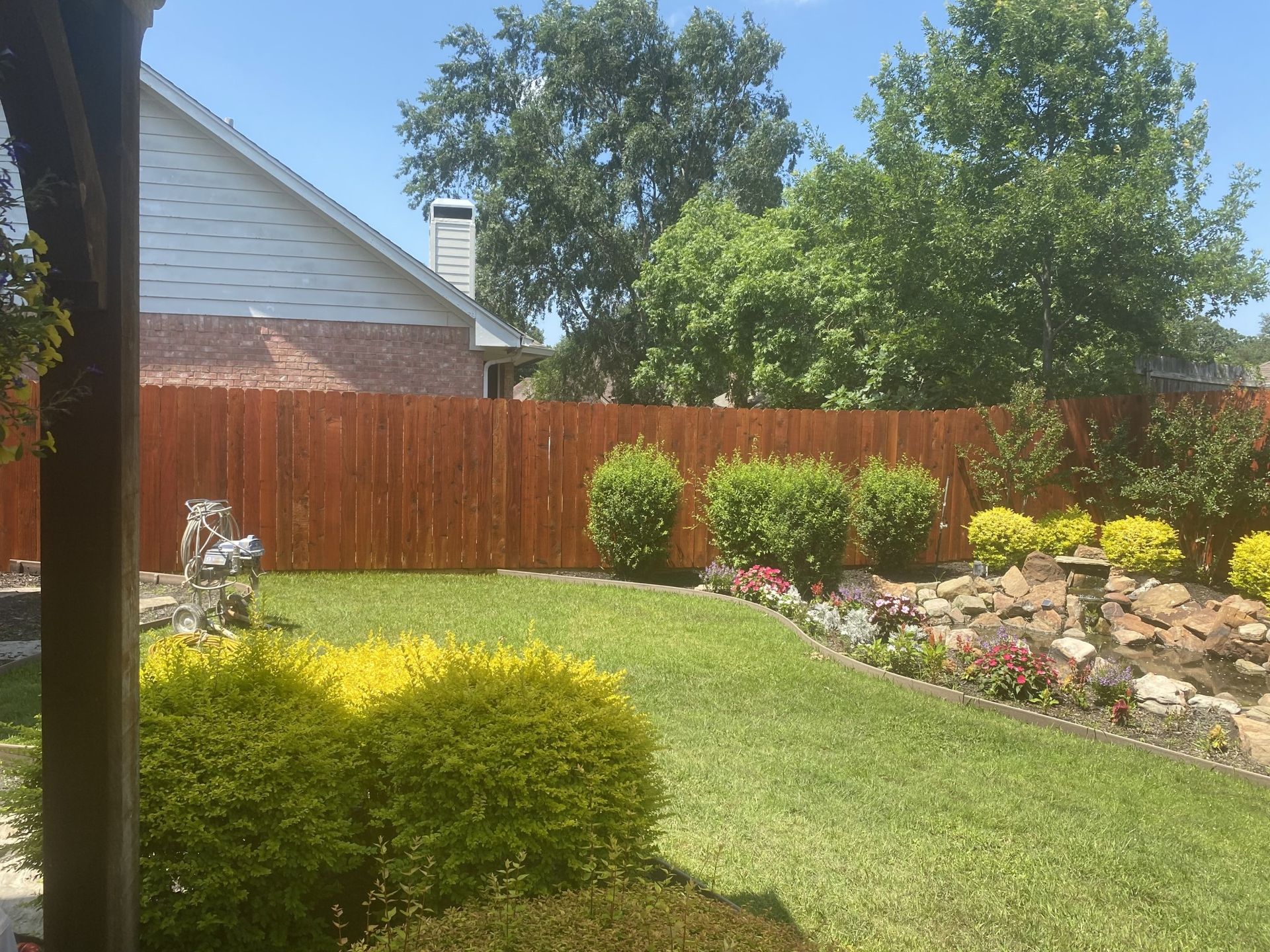Lush green backyard with a stained wooden fence, bushes, and a small rock water feature.