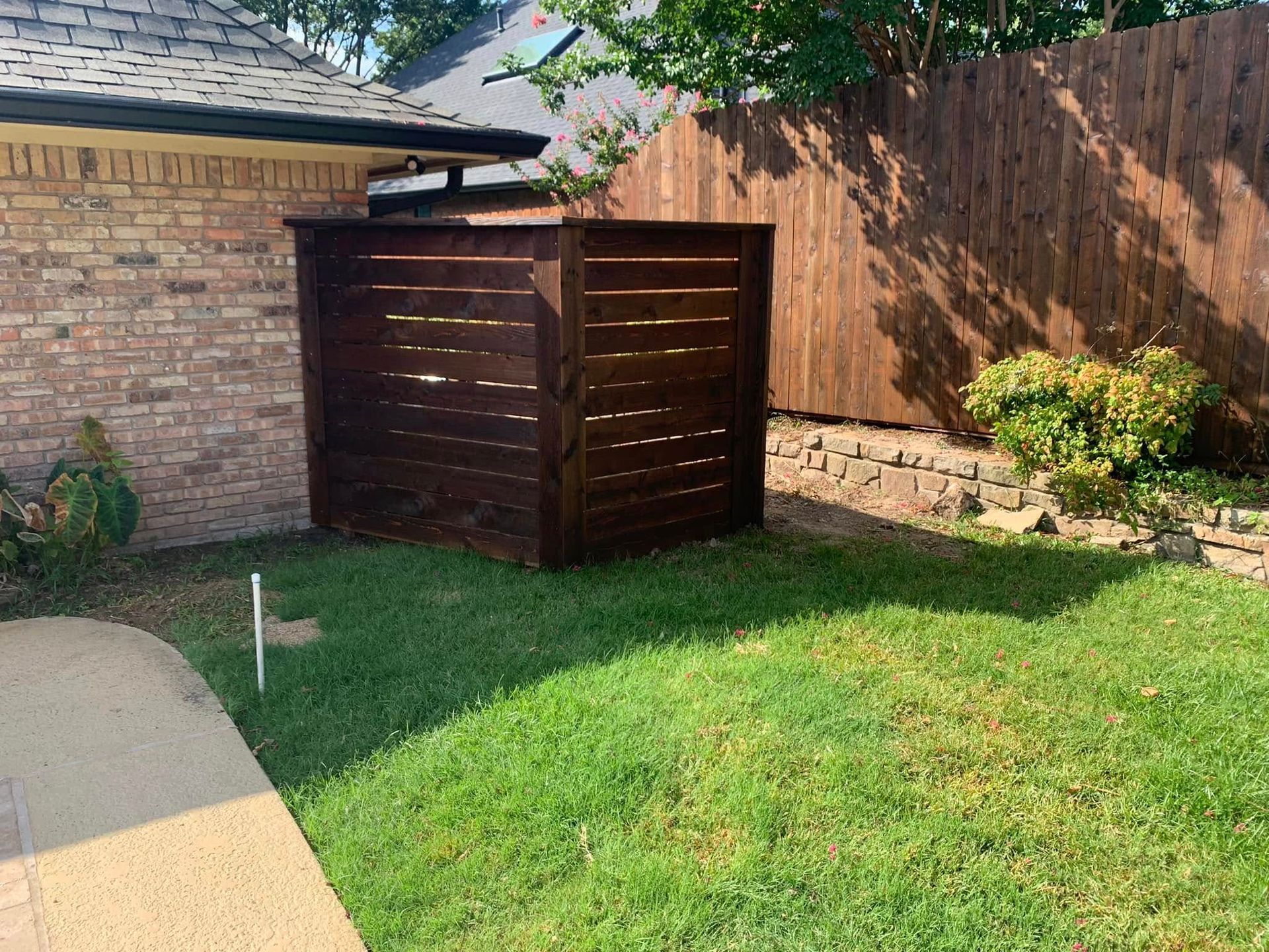 Brown wooden trash enclosure next to a brick house, on green grass in a yard.