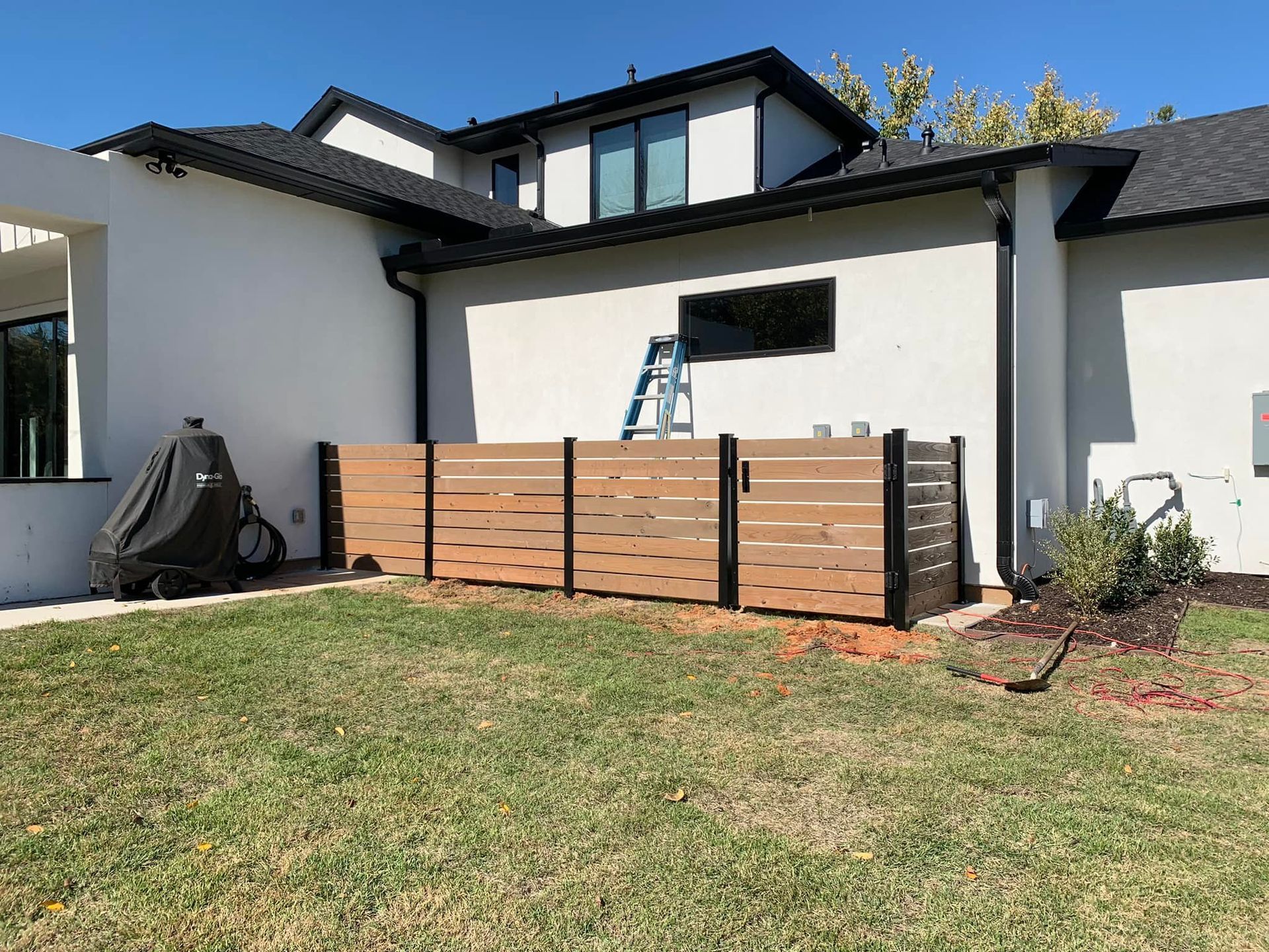 Backyard with a brown and black wooden fence, lawn, and a white house.