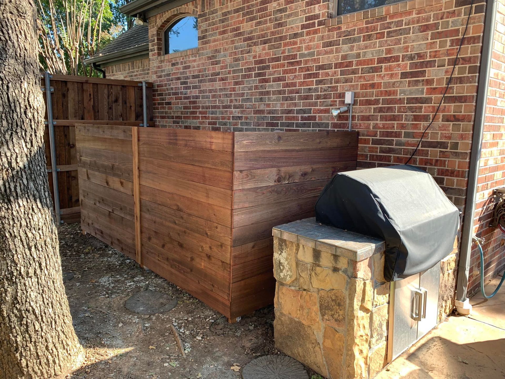 A brick house exterior with a wooden privacy fence and grill on a stone base.