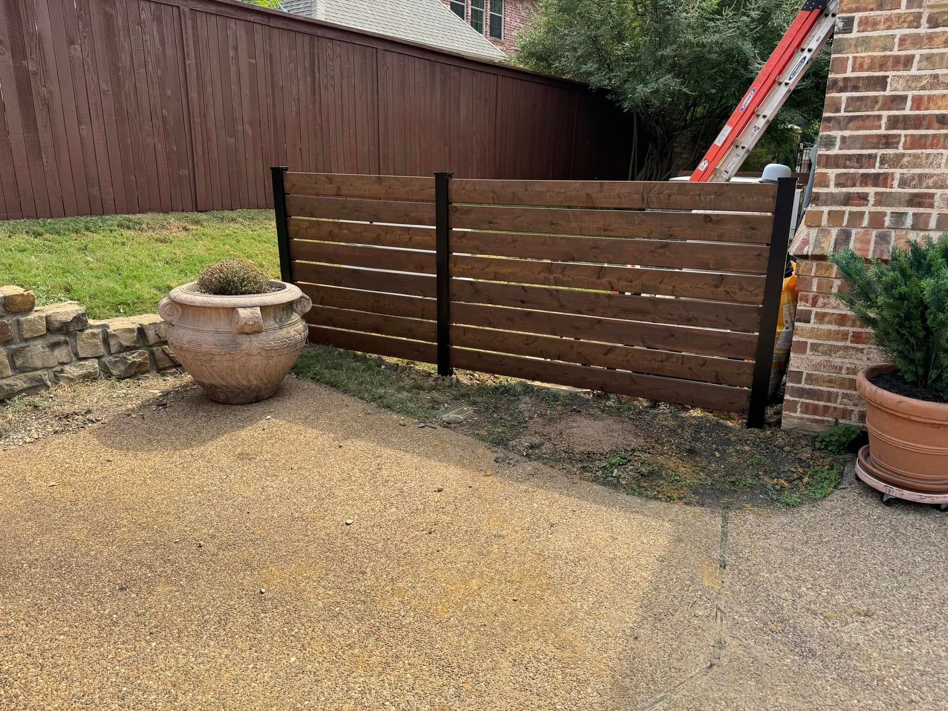 Brown wooden fence section with black posts on gravel patio, plants on either side.