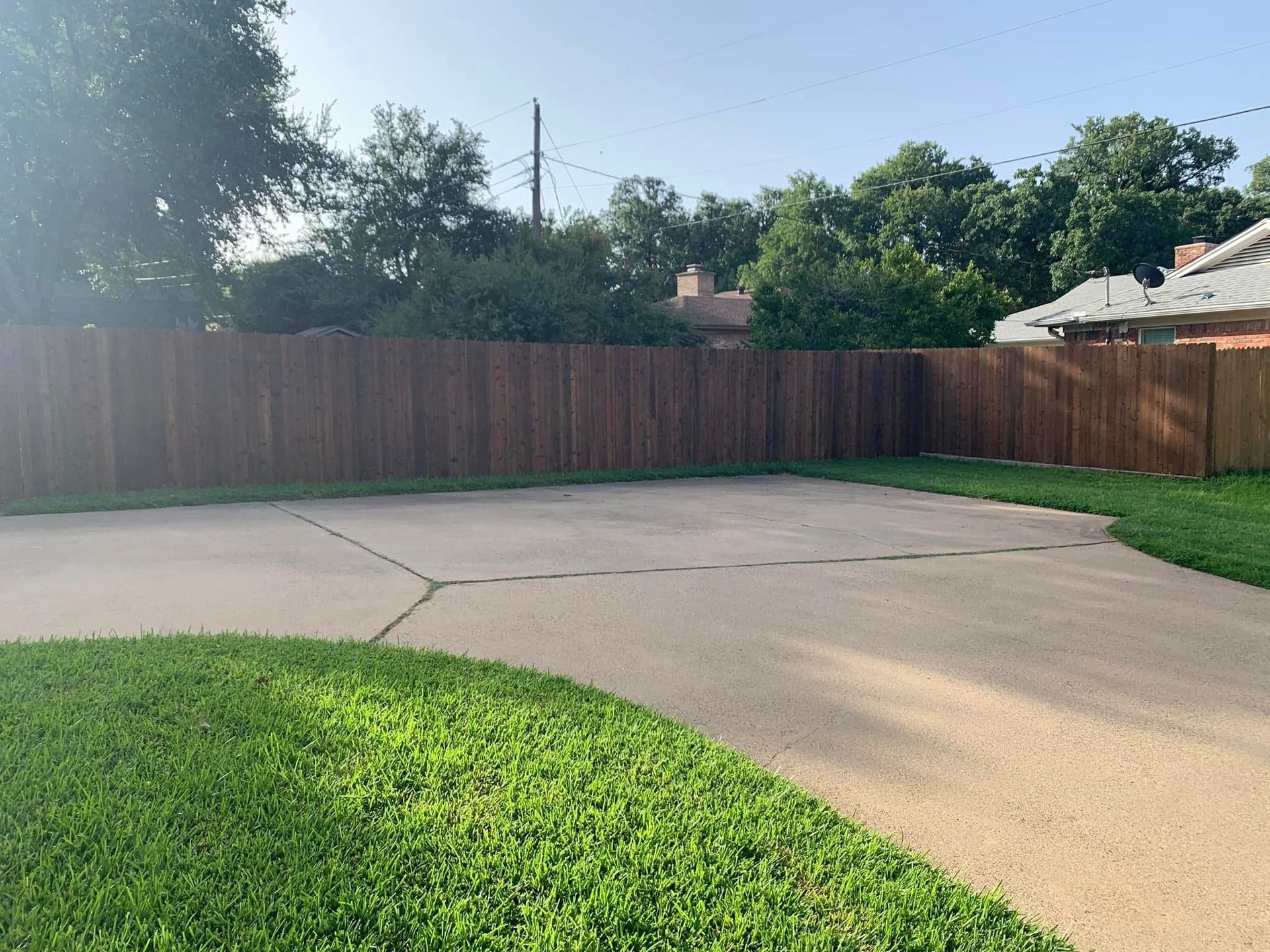 A concrete driveway bordered by green grass and a stained wooden fence under a sunny sky.